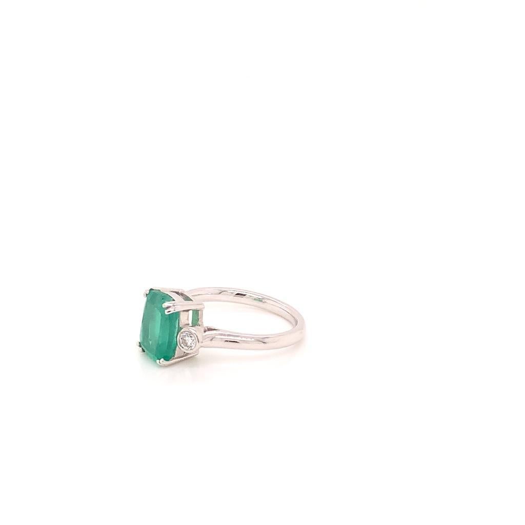 The sparkling cushion cut emerald at the centre of this unique ring weighs approximately 2.56 carats and is claw set in 18k white gold. The diamonds on either side of this precious stone which are F/G colour, SI clarity, and weigh approximately 0.40