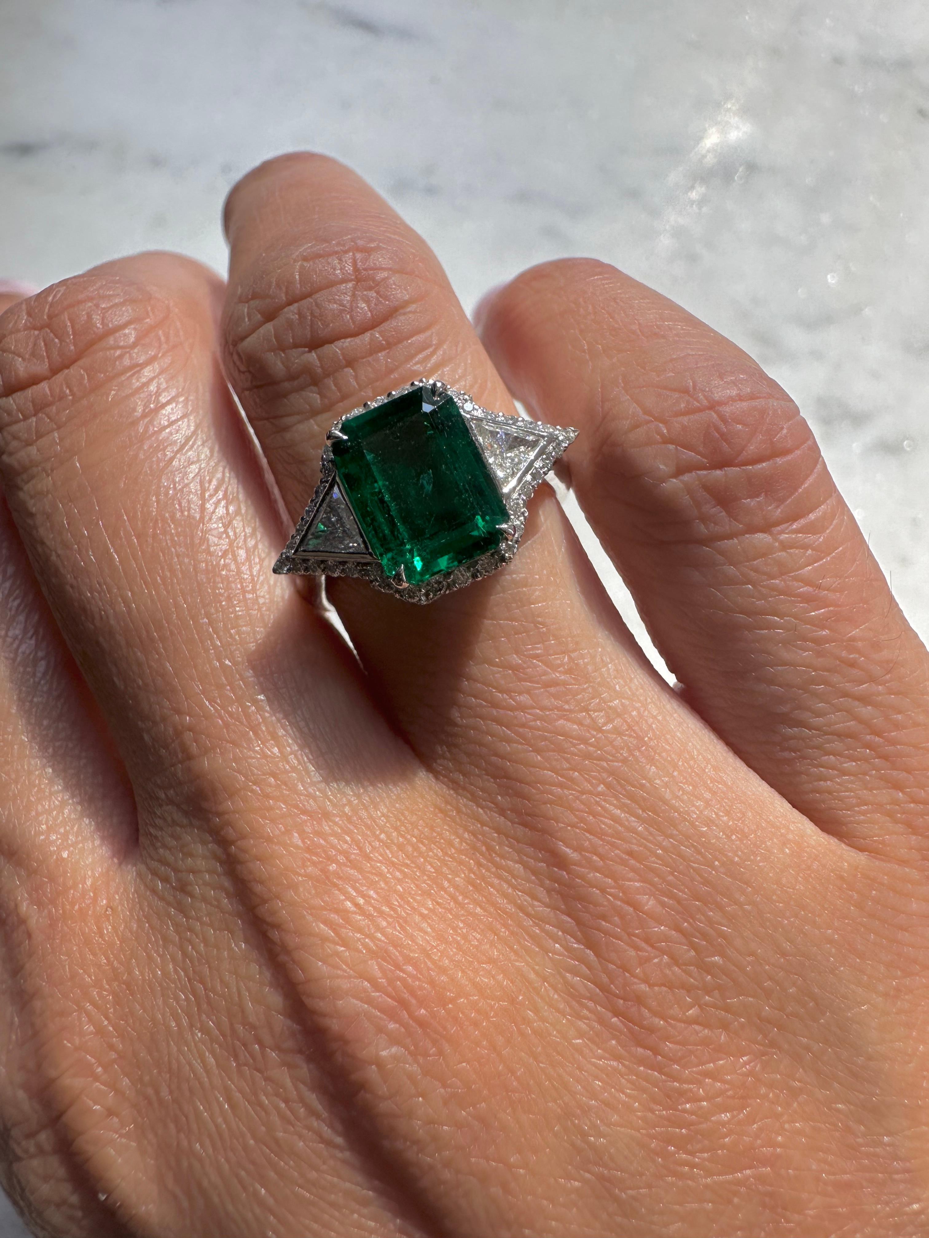This unique ring is Art Deco inspired, while maintaining a strikingly modern feel.
 
The 2.57 carat Zambian emerald has a vibrant green color. It is low oil certified by AGL.

The emerald is set between two GH VS+ trillion diamonds. The entire shape