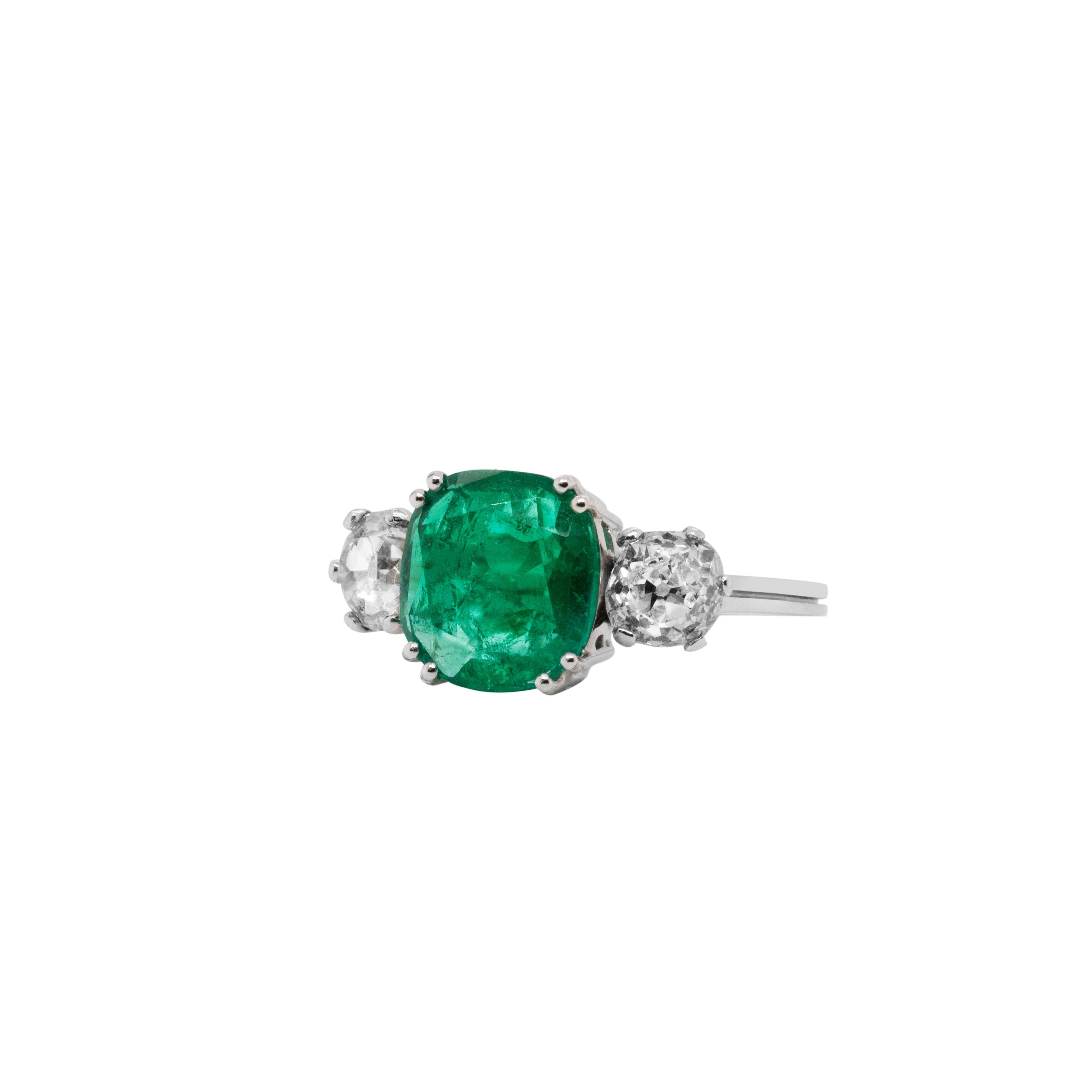 This gorgeous vintage three-stone engagement ring features a rectangular cushion cut Colombian emerald weighing 2.58 carats, mounted in a four double claw, open back setting. The vibrant green gemstone is beautifully accompanied by a Victorian old