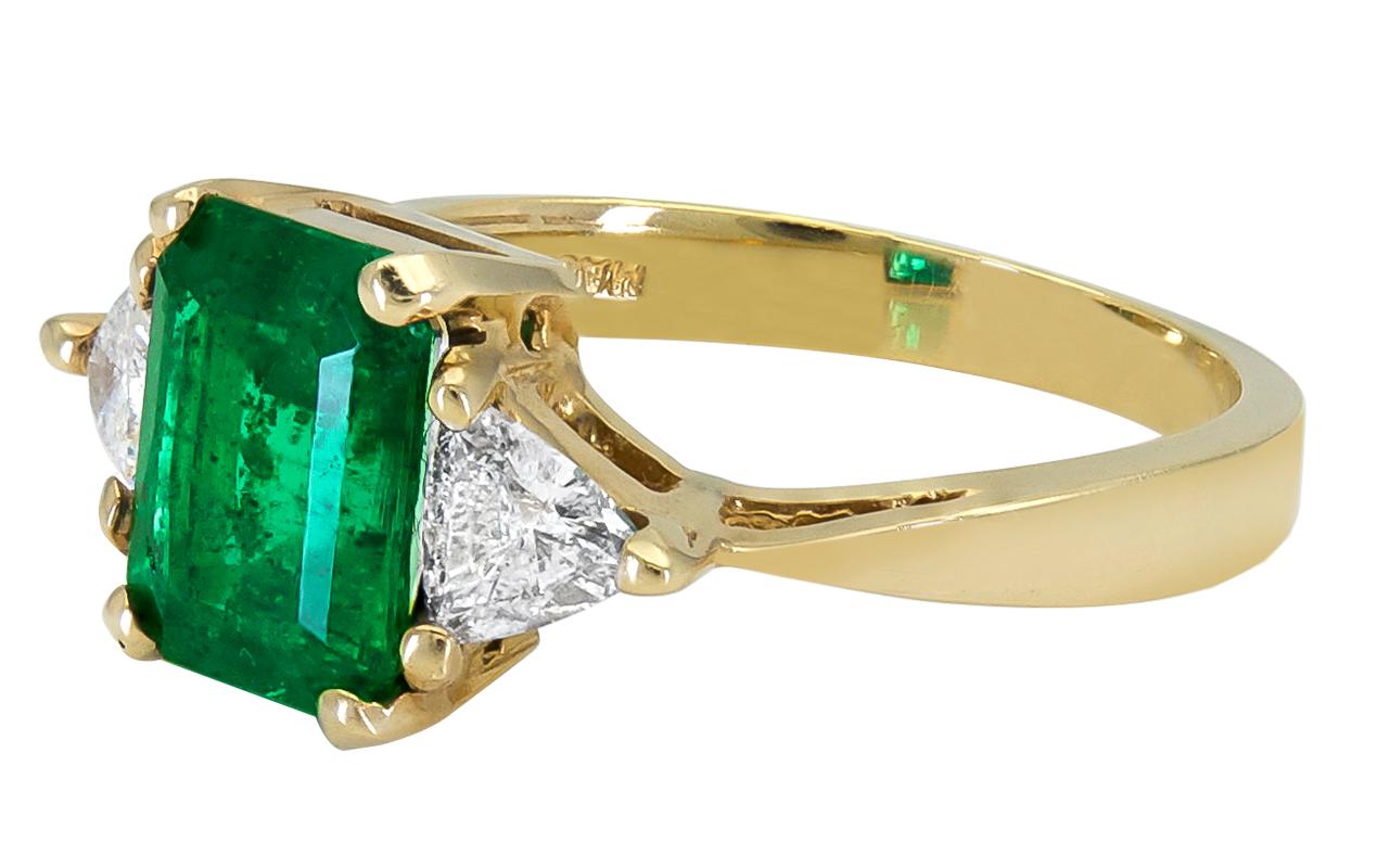 A classic three-stone engagement ring showcasing a color-rich emerald cut green emerald, accented with brilliant trillion (triangular) diamonds on either side. Set in a tapered 18k yellow gold mounting. 
Emerald weighs 2.59 carats.
Diamonds weigh