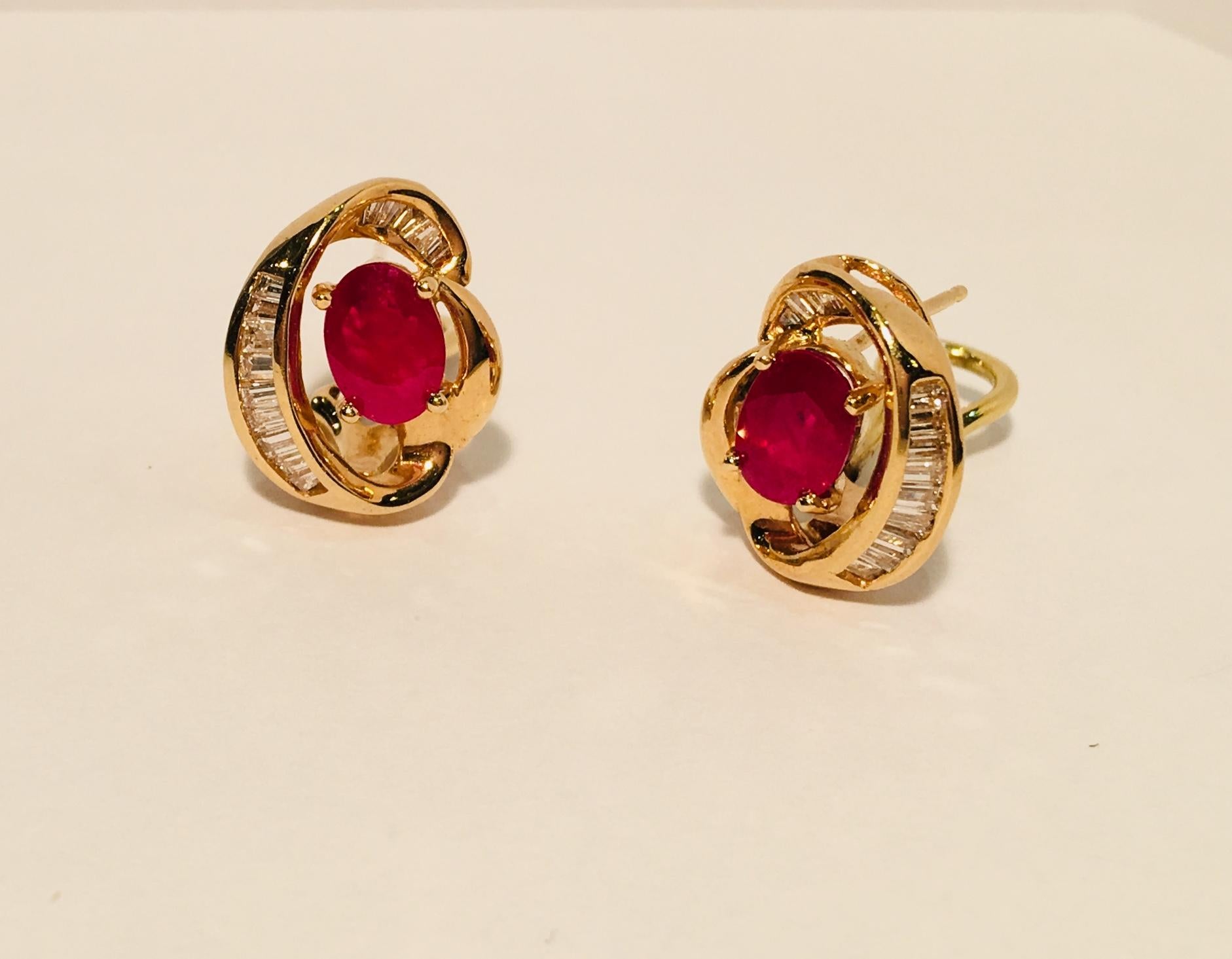 Classic 14 karat yellow gold omega or lever back earrings for pierced ears feature a prong set, oval cut, bright pigeon blood red ruby surrounded by swirls of invisibly set baguette diamonds.

2 rubies measure 7.19 mm x 5.09 mm x 3.4 mm and weigh