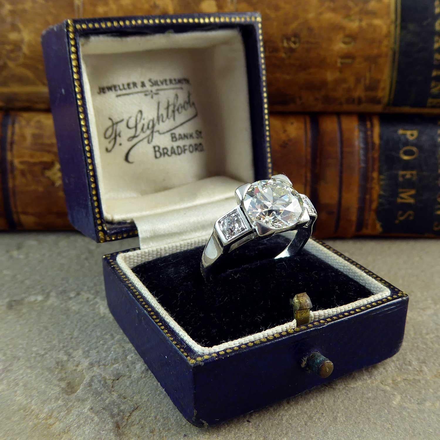 A fabulous diamond solitaire engagement ring featuring a four claw set centre early round brilliant cut diamond in four claw setting to a white square basket mount to square profile shoulders each set with an old European cut diamond  The diamond