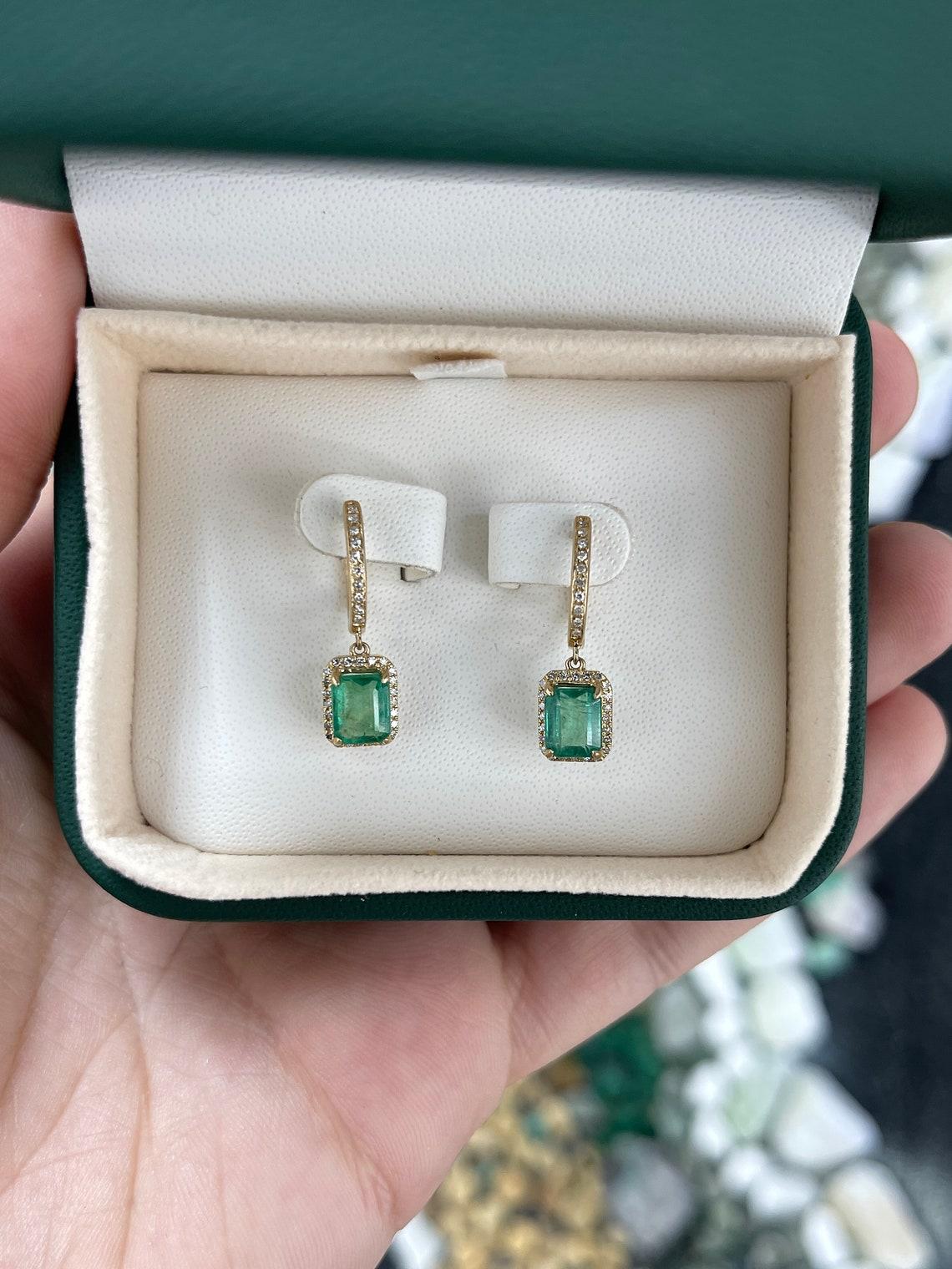 Indulge in the epitome of luxury with our stunning emerald and diamond dangle earrings. Exuding opulence, these earrings boast a total of 2.25-carats of lush green emerald cut emeralds, elegantly prong-set and adorned with a radiant diamond halo.
