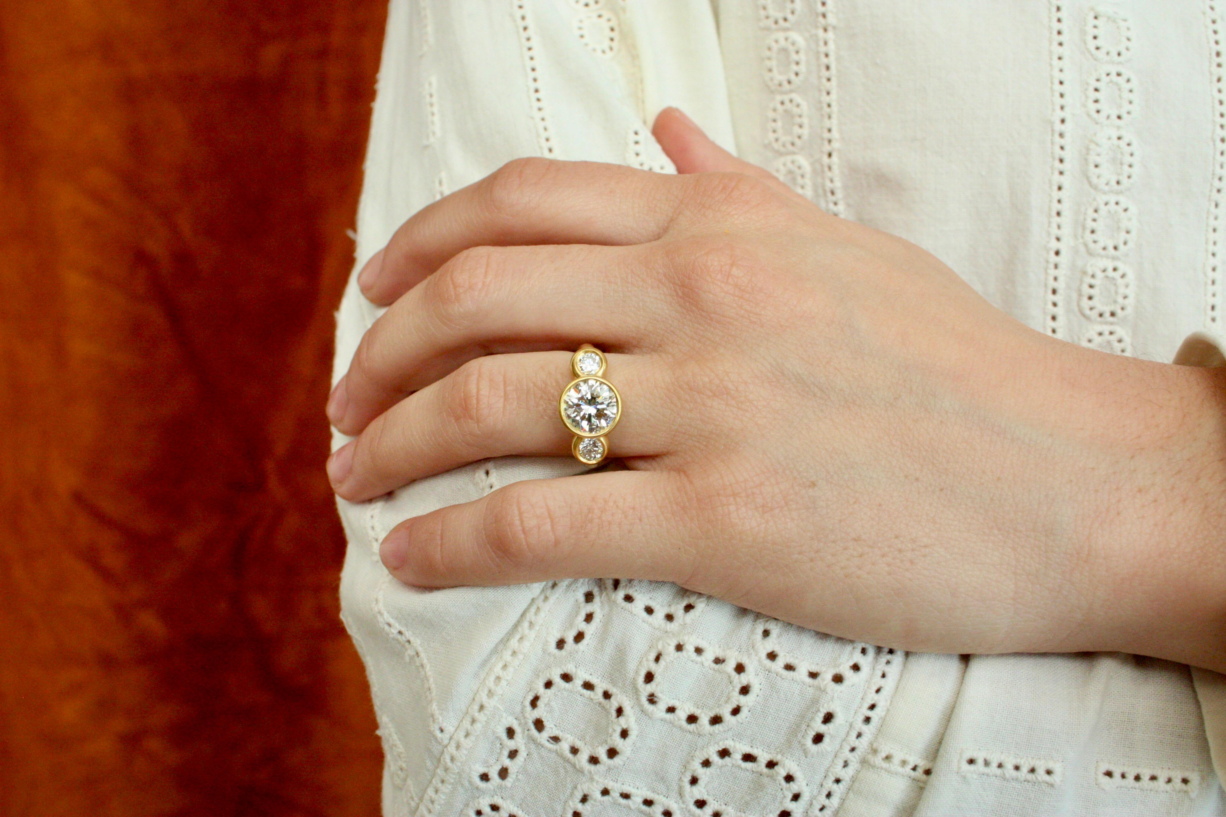 This breathtaking engagement 3 stone ring features three round-cut diamonds totaling 2.65 carats, set in a rich 22k yellow gold bezel setting with a soft-hammered finish. The centerpiece, a 2.15ct natural diamond, is complemented by two smaller