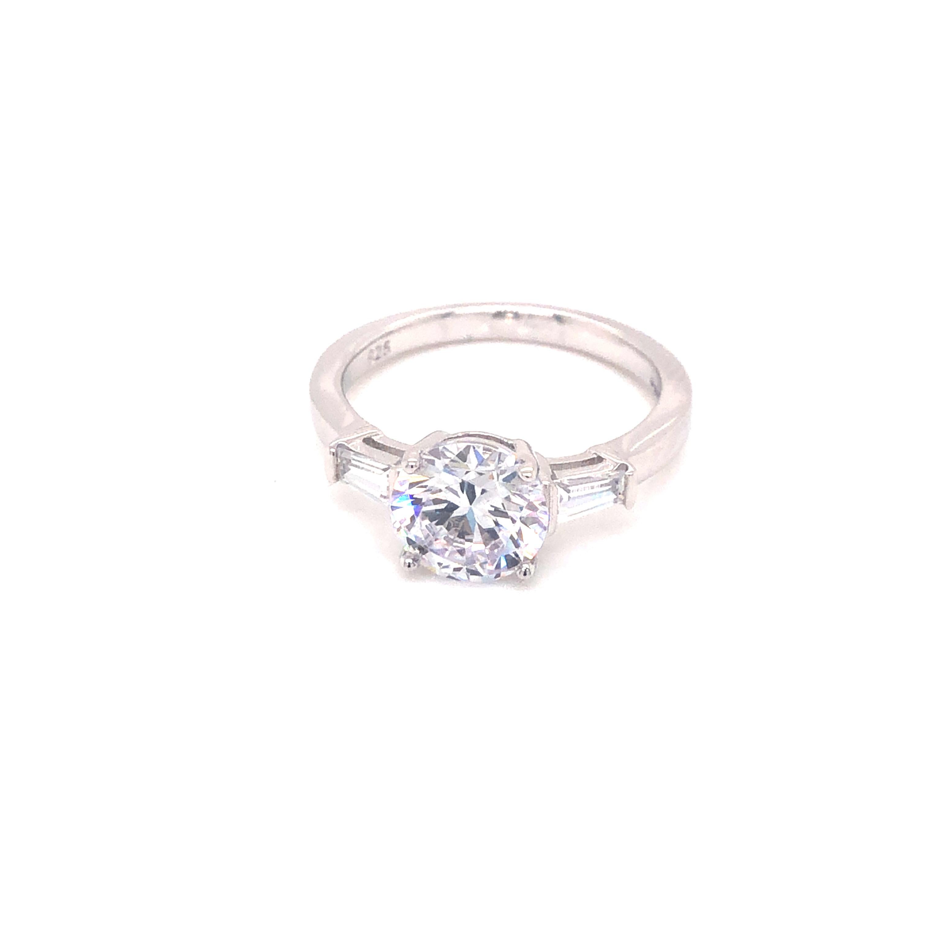 This elegant trilogy style ring features a central round brilliant cut measuring 2.69 carat cubic zirconia flanked by two matching tapered baguettes measuring 0.30 carat each. 

Composed of 925 sterling silver with a high gloss white rhodium