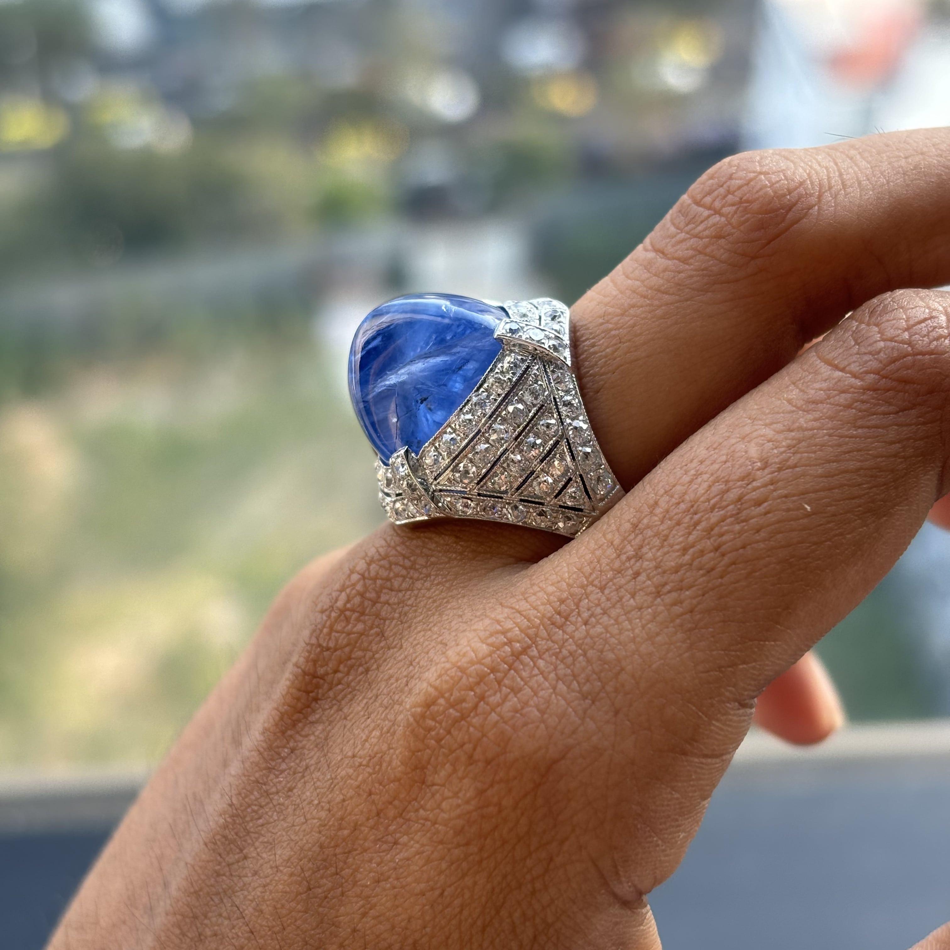 Prepare to be enthralled by the captivating allure of this masterpiece that beckons you to immerse yourself in a world of timeless beauty and luxury. At its heart lies a resplendent 27.05 Carat Sugarloaf Sapphire, a gem of unparalleled magnificence