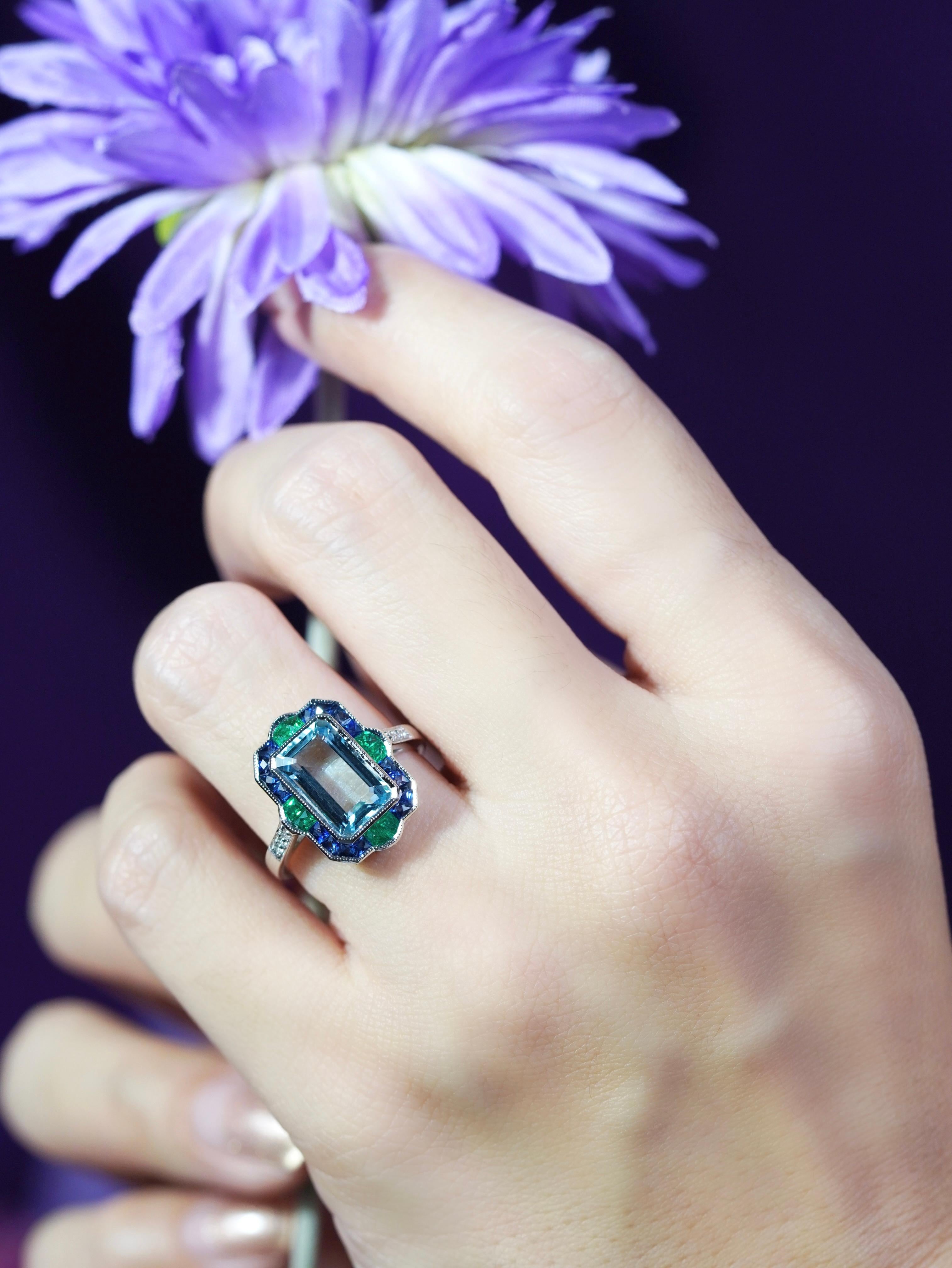 Au centre, une aigue-marine de 2,76 carats, taillée en émeraude. La pierre bleue sereine est entourée d'un halo de riches saphirs taillés à la française, créant un motif cible saisissant. Quatre émeraudes vives accentuent chaque coin - en haut, en