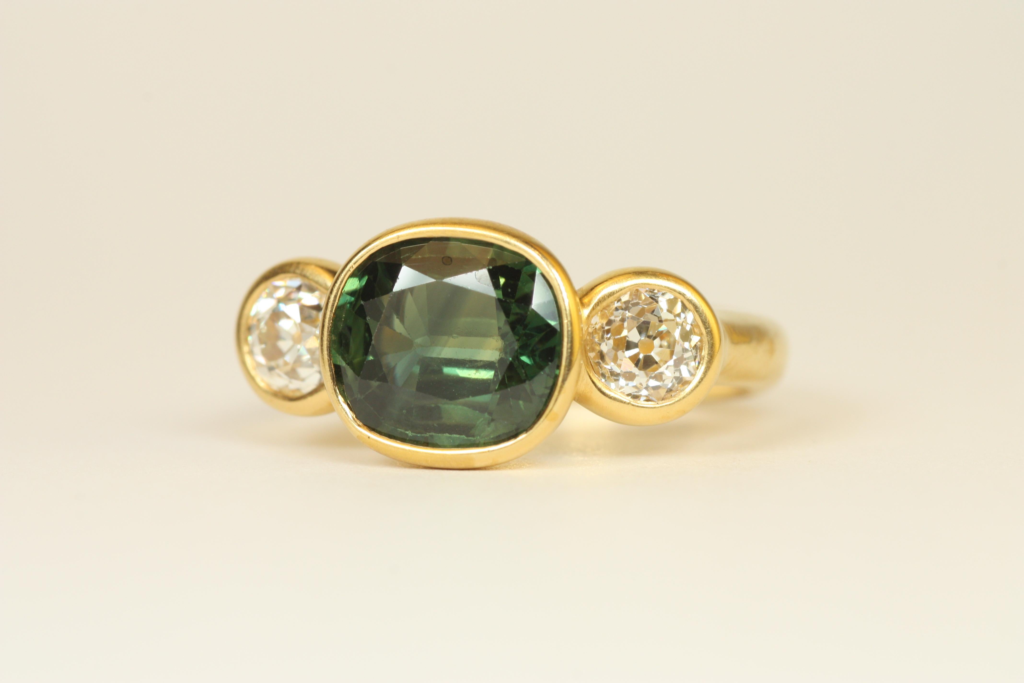 Cette bague distinctive à trois pierres est ornée d'un saphir vert ovale de 2,78ct, flanqué de deux diamants taillés dans des mines anciennes pour plus d'éclat. Sertie dans de l'or jaune martelé 22k, cette pièce unique allie l'élégance ancienne à