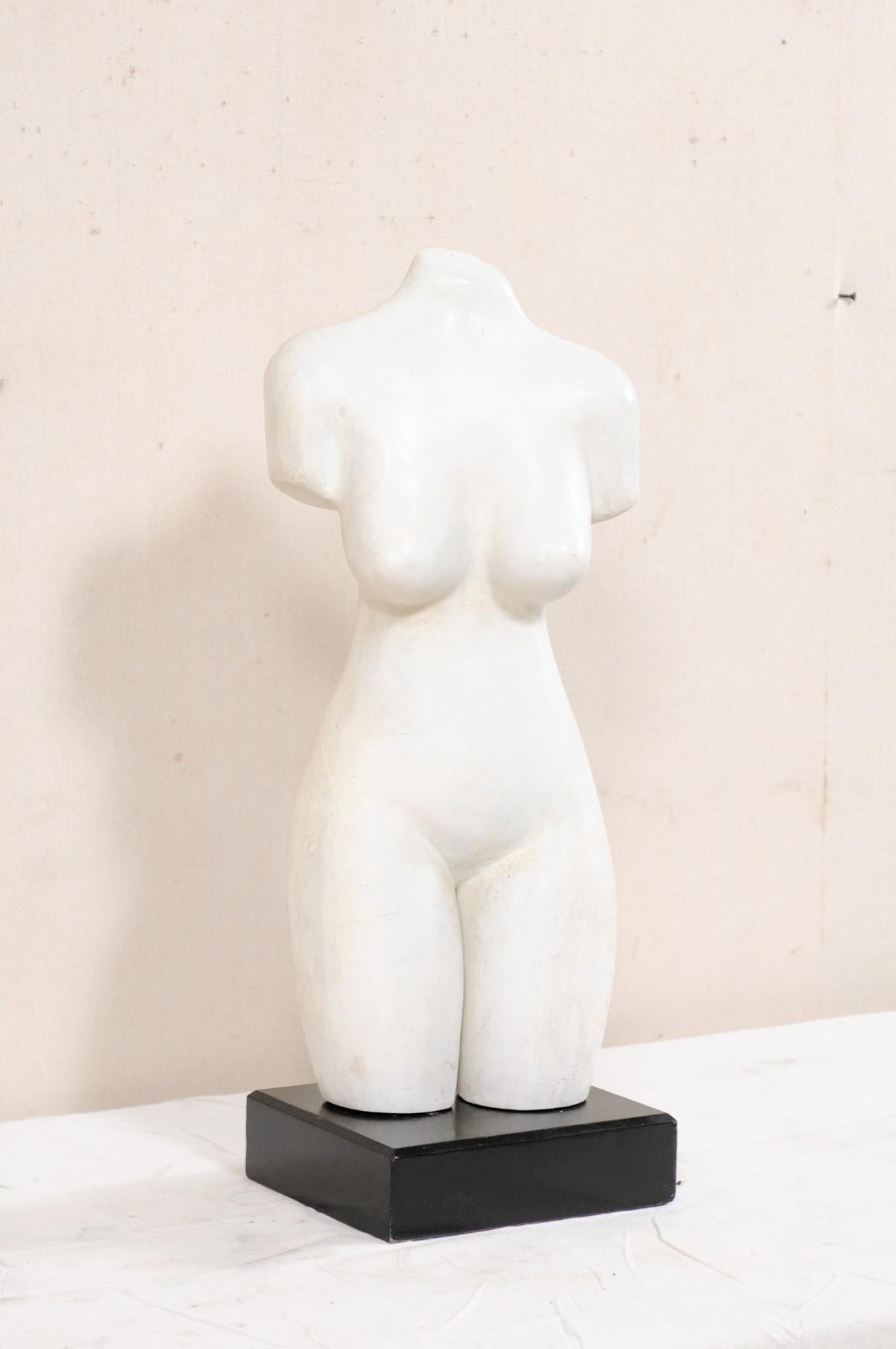 An artistic nude female torso statue on stand. This vintage American composite sculpture depicts a nicely cured torso, or female's mid region, raised on a square-shaped base. This artisan feminine piece from the 20th century is done 