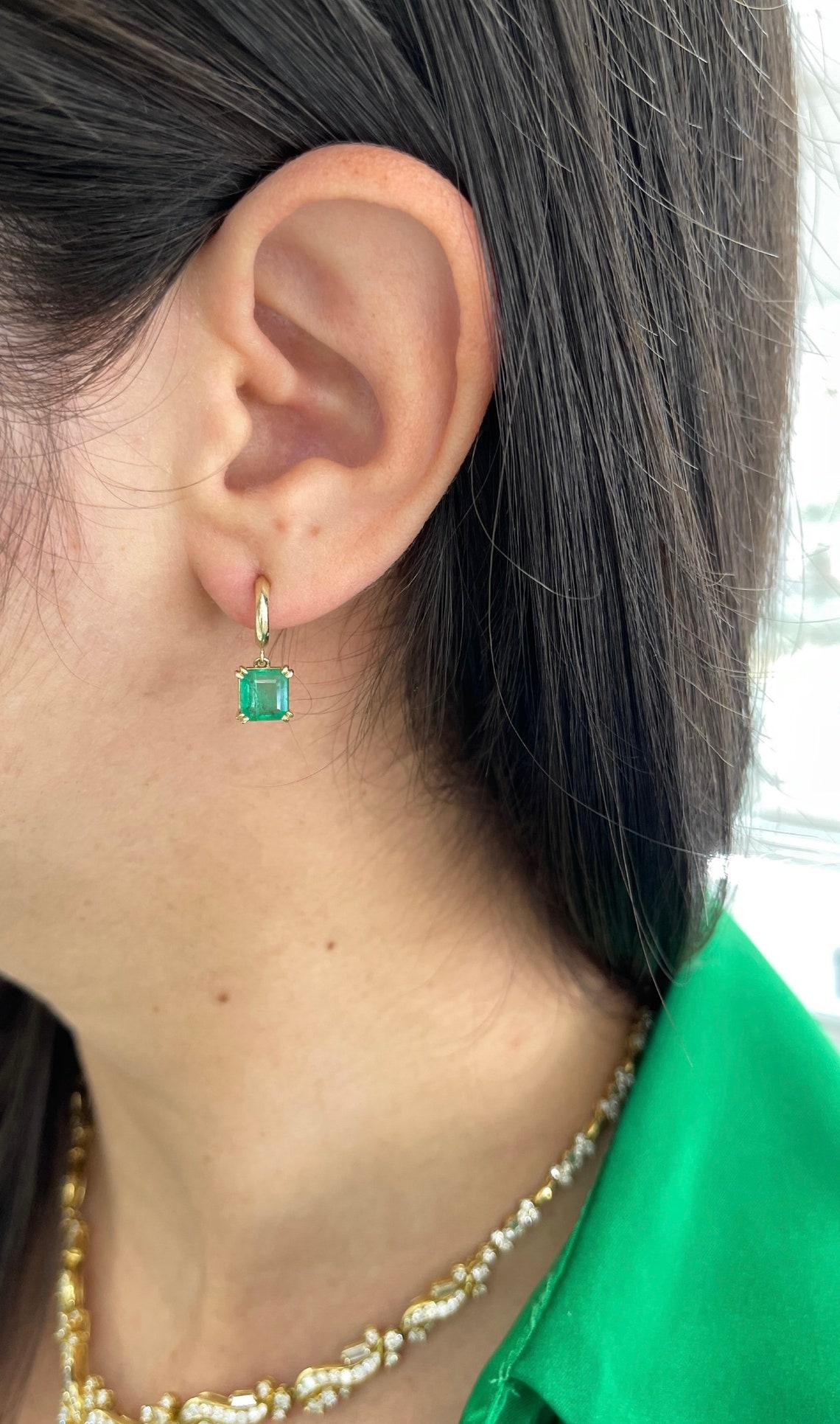 These emerald dangle earrings feature two mesmerizing asscher cut natural emeralds, displaying a medium dark green color with good clarity and luster. The emeralds are securely set in a double claw prong setting, highlighting their unique shape and