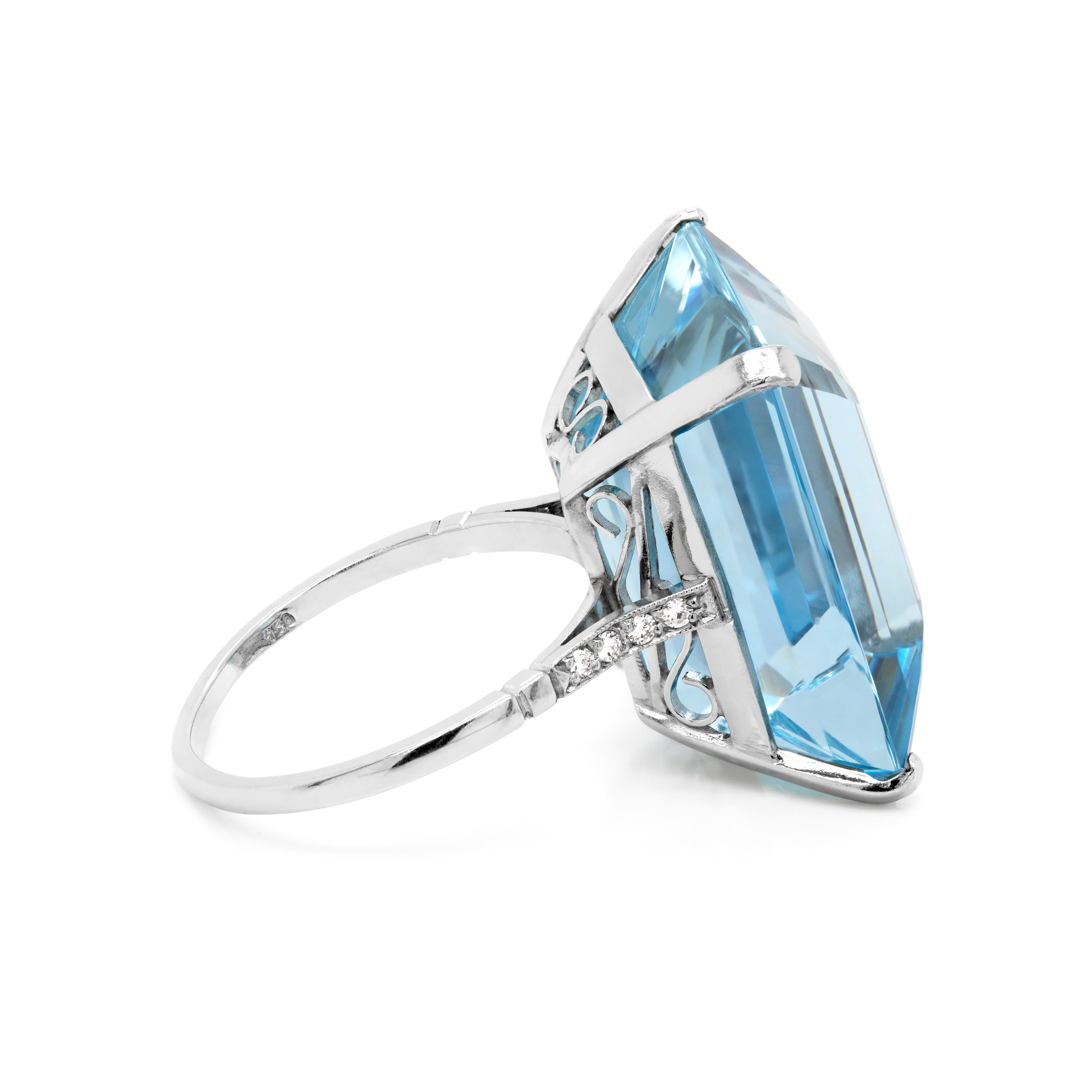 This remarkable Art Deco ring beautifully features an emerald cut aquamarine weighing an impressive 28.50 carats mounted in a four claw, open back setting. The gemstone is sat in an openwork gallery with delicate scrolling motifs across the collet.