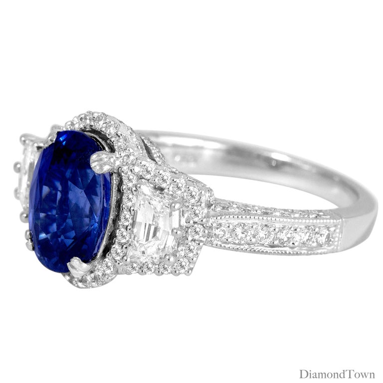 This beautiful ring has a 2.97 carat oval cut Ceylon Sapphire center, flanked by two cadillac cut diamonds. The center sapphire and the two main diamonds are each surrounded by a halo of round diamonds, which also extends down the side shank. The