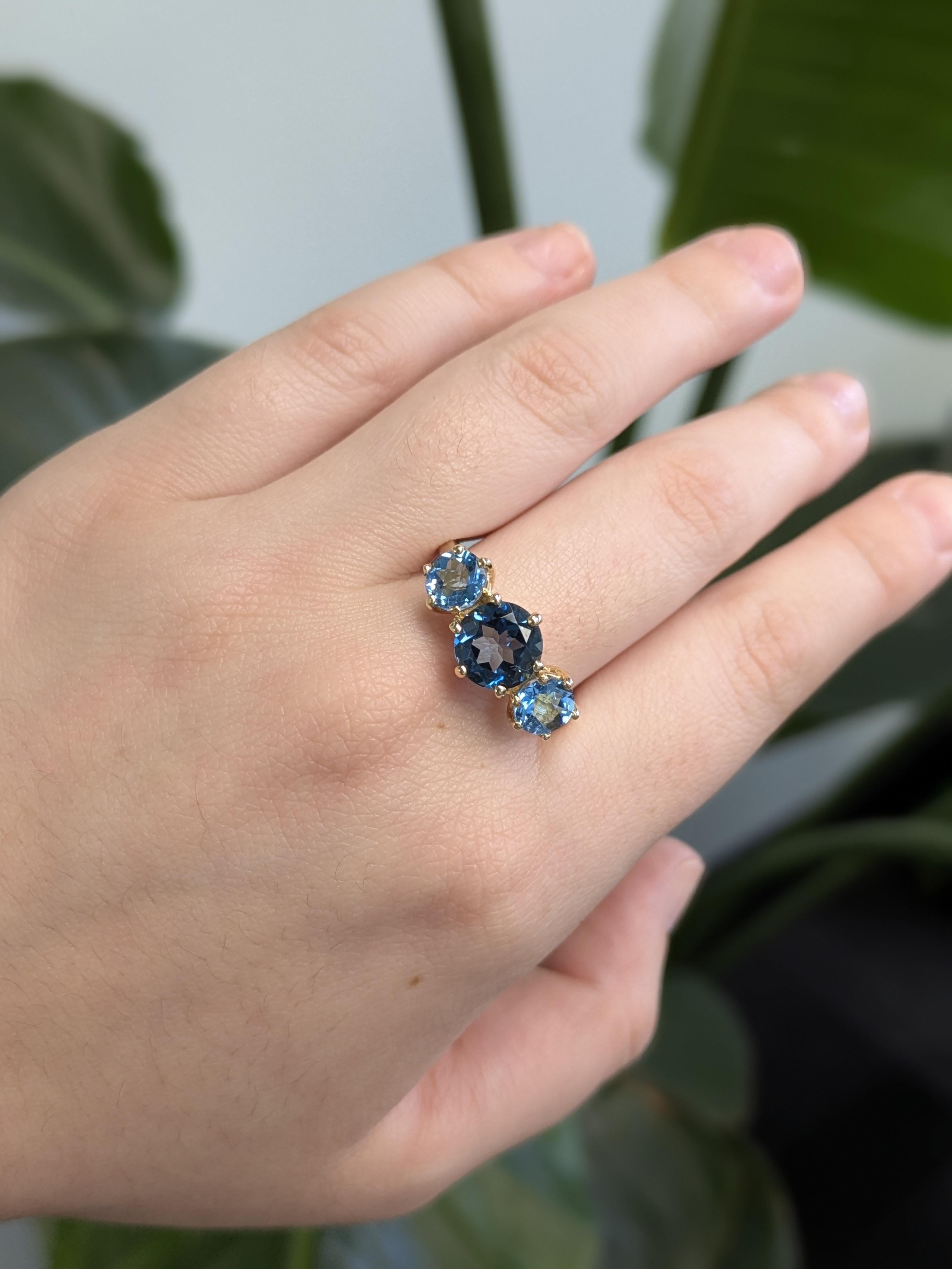 Elegante anello a tre pietre con un topazio blu London rotondo al centro, affiancato da due pietre d'accento scintillanti. Realizzato in oro giallo massiccio 14 carati, questo design senza tempo irradia un colore ricco e una raffinatezza