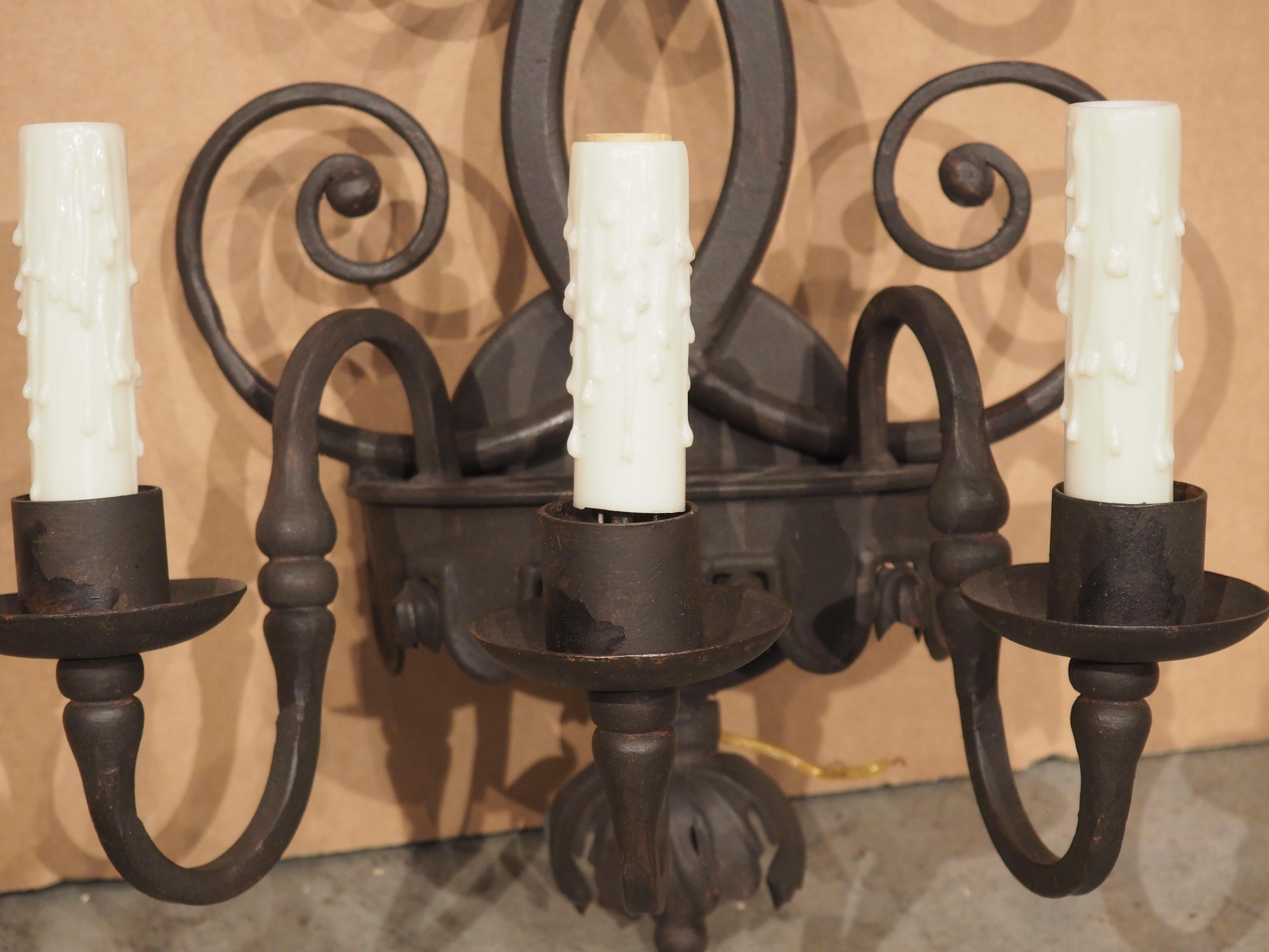 Dieser dreiarmige Wandleuchter aus Eisen ist im Stil spanischer Architekturbeleuchtung geschmiedet und wird von weißen, künstlichen Kerzenhülsen gekrönt, die aus Untertassen herausragen. Jeder Arm hat eine gewundene S-Rollenform mit einem gedrehten