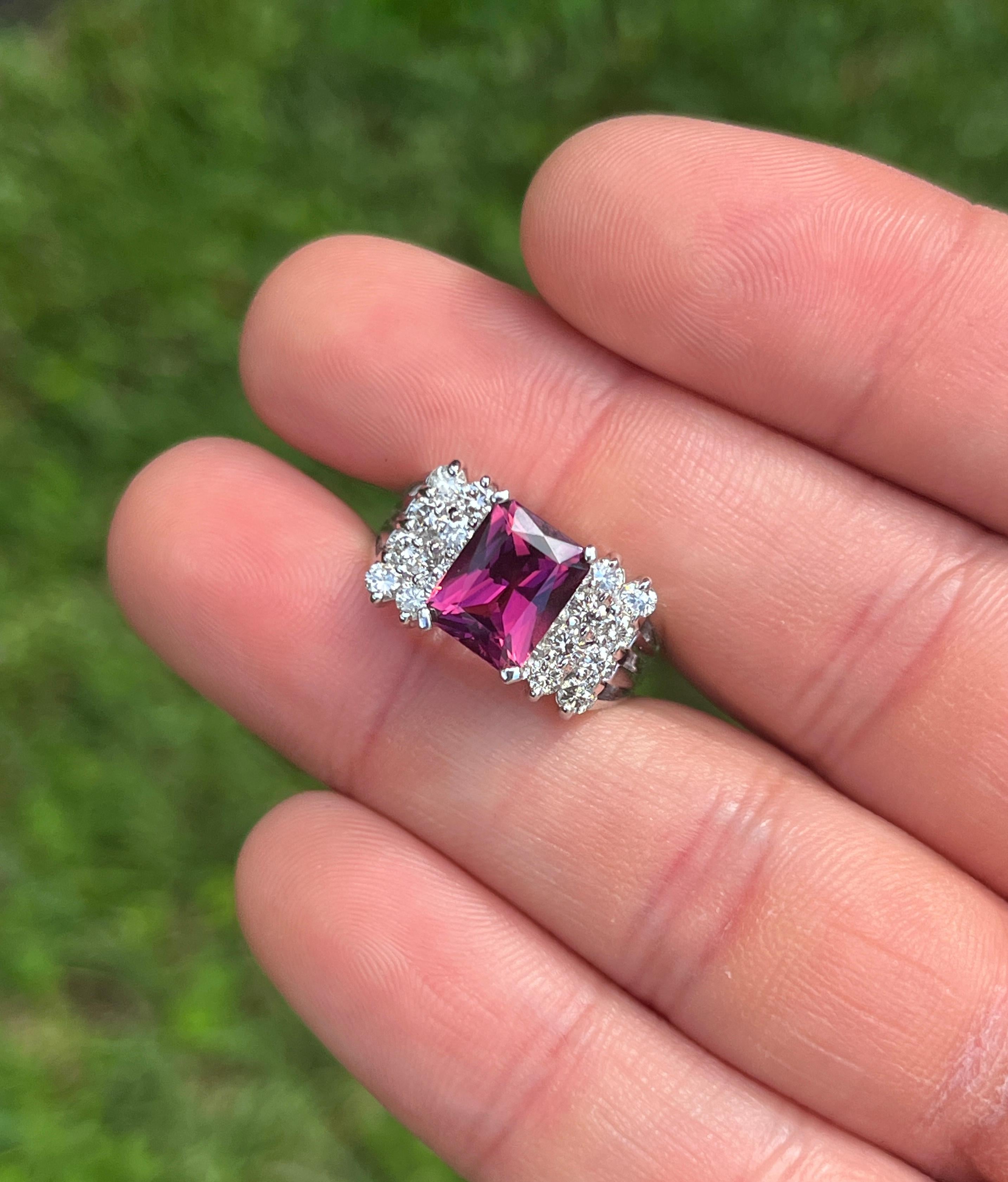 Vintage 3 Carat Radiant Cut Vivid Pinkish Purple Tourmaline Ring. Set in Platinum 950, this semi-precious gemstone ring weighs 9.6 grams and exudes the charm of a vintage era.

Flanked by a cluster of round-cut white diamonds and securely 4-prong