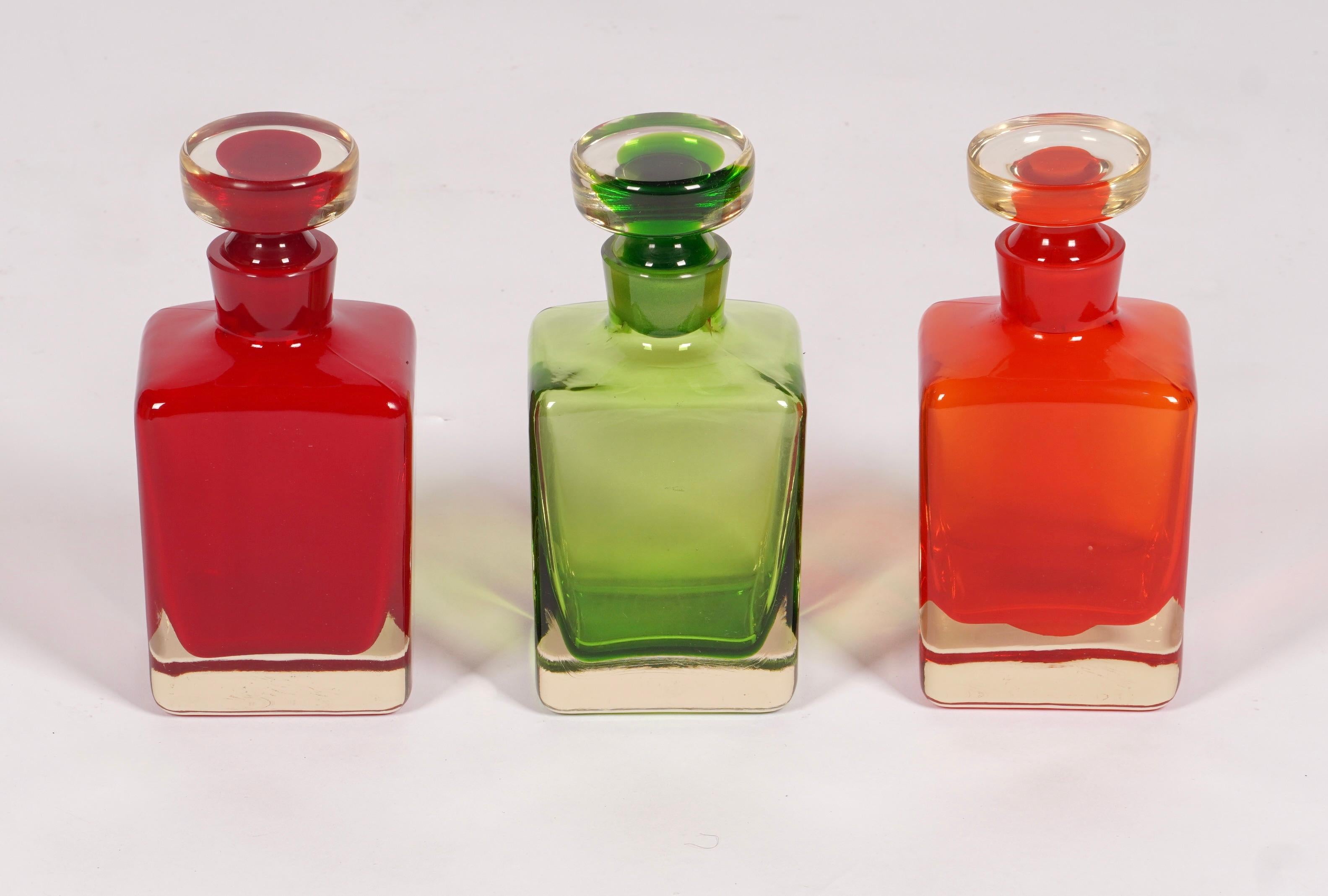 3 farbenfrohe Glaskaraffen in Grün und zwei Rottönen, eine in einem dunkleren Rot und die andere mit Orange in i. Rechteckige Körper mit spitz zulaufenden, runden Stopfen. Die Farben haben eine durchscheinende Qualität, besonders wenn sie von hinten