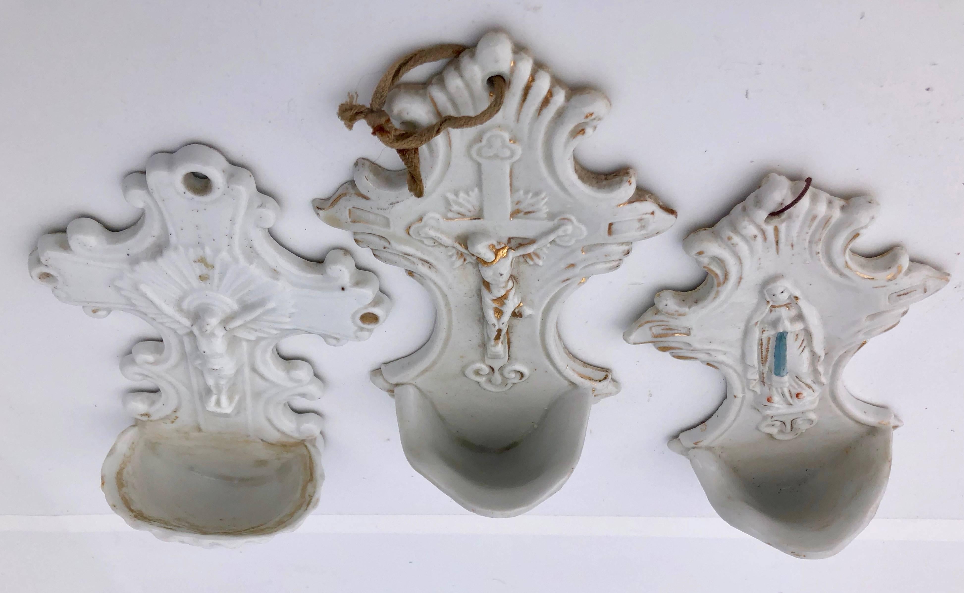 This is a wonderful collection of French antique holy water fonts (benitiers). There are two white porcelain holy water fonts shaped like a cross with crucifixes (one has gold on it) and one with Mary providing a blessing, which has some blue and