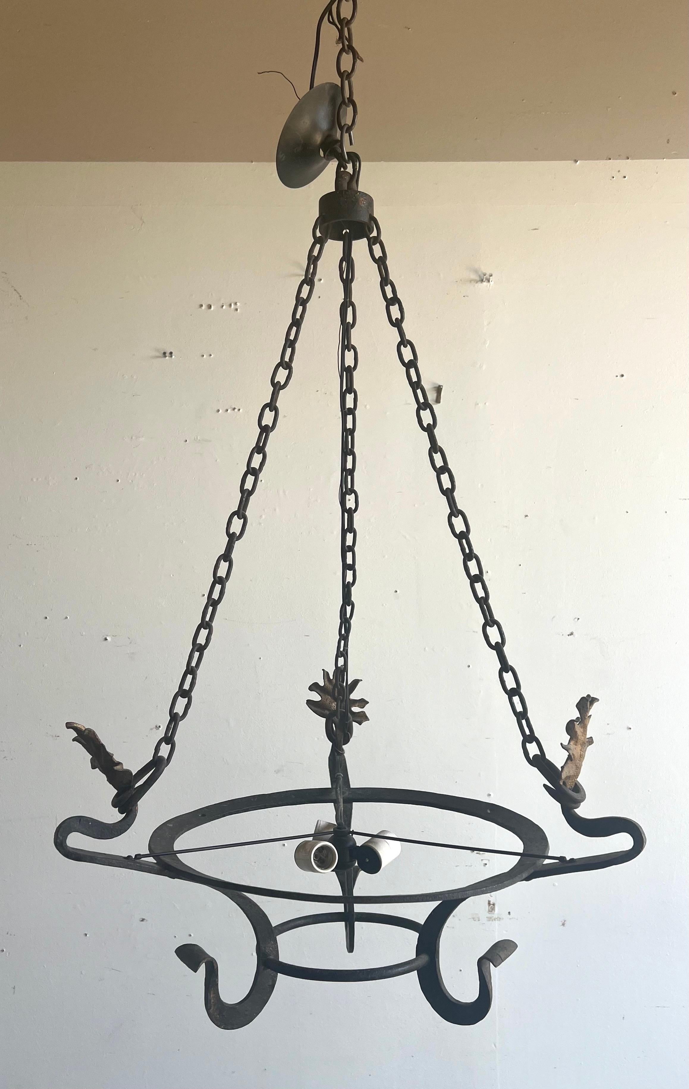 Lustre industriel minimaliste de style espagnol présentant un design simple à cadre ouvert, avec un anneau central soutenu par trois chaînes reliées à une monture de plafond.  L'armature métallique est en fer forgé foncé, ornée de feuilles d'acanthe