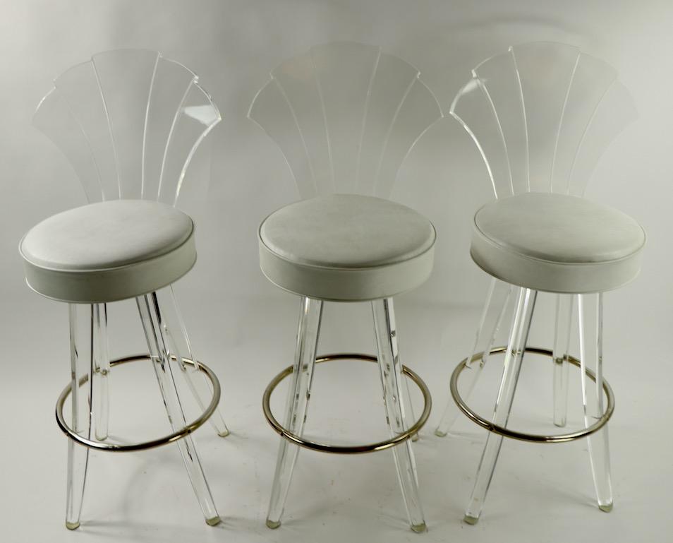 Nice set of three counter height fan back cast Lucite, chrome and upholstery stools. The stools have Art Deco revival stepped form fan backrests, off white vinyl upholstered pad seats, Lucite legs and a chrome ring footrest. Measures: Pad seat 15