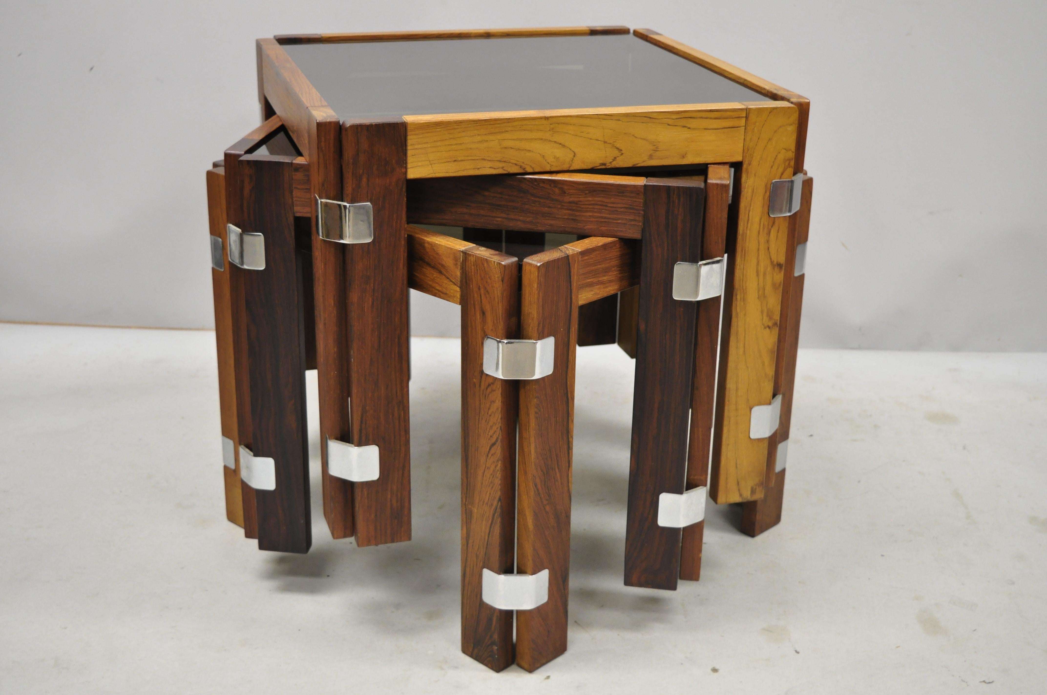 Set of 3 midcentury Danish modern rosewood and smoked glass side tables by interior form. Listing includes polished metal joinery, nesting form, smoked glass tops, solid wood construction, beautiful wood grain, original label, clean modernist lines,