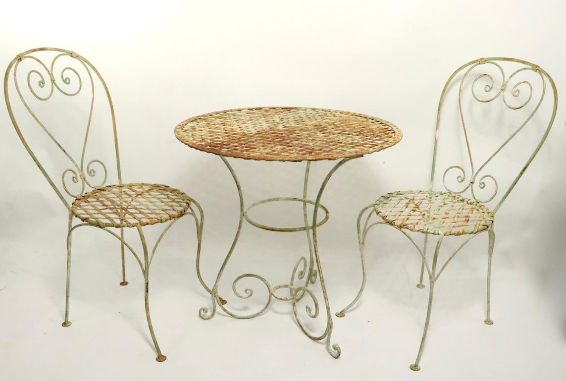 Charming 3 piece bistro set, consisting of two side chairs and a cafe style dinette table. Woven iron seats and table top, curvilinear frames, patinated old paint and light rust surface. The table top has a hand riveted edge which adds to the design