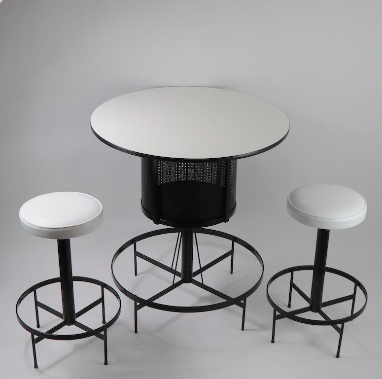 Very chic and stylish bar and stool set comprised of 2 stools and a round bar. The bar has a mesh enclosed storage space, which has a curved sliding door, under the circular white Formica top. The stools have newly upholstered white vinyl pad seats,
