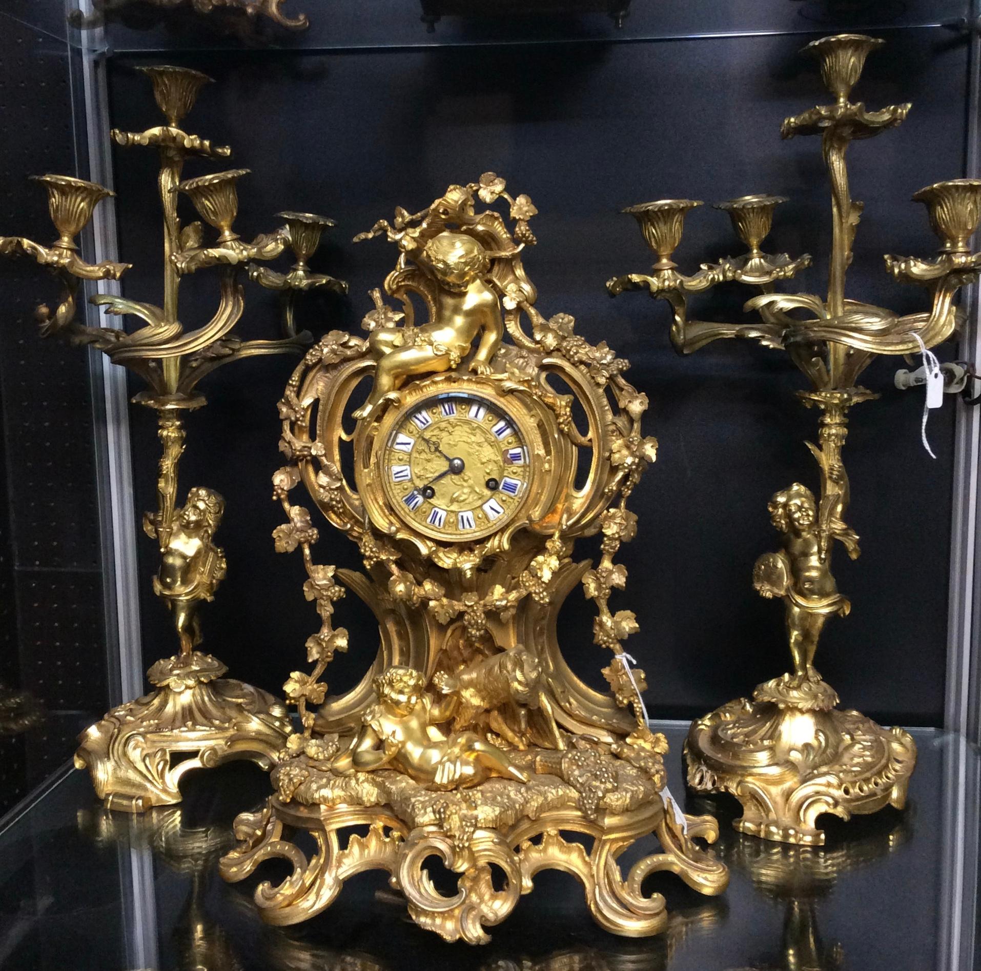 A beautiful three piece bronze gilt clock set, this lovely piece has been ornately decorated with gold leaves and cherubs. Its bronze face has a telephone style dial on enamel and blue roman numerals, the clock is striking on a bell and comes with