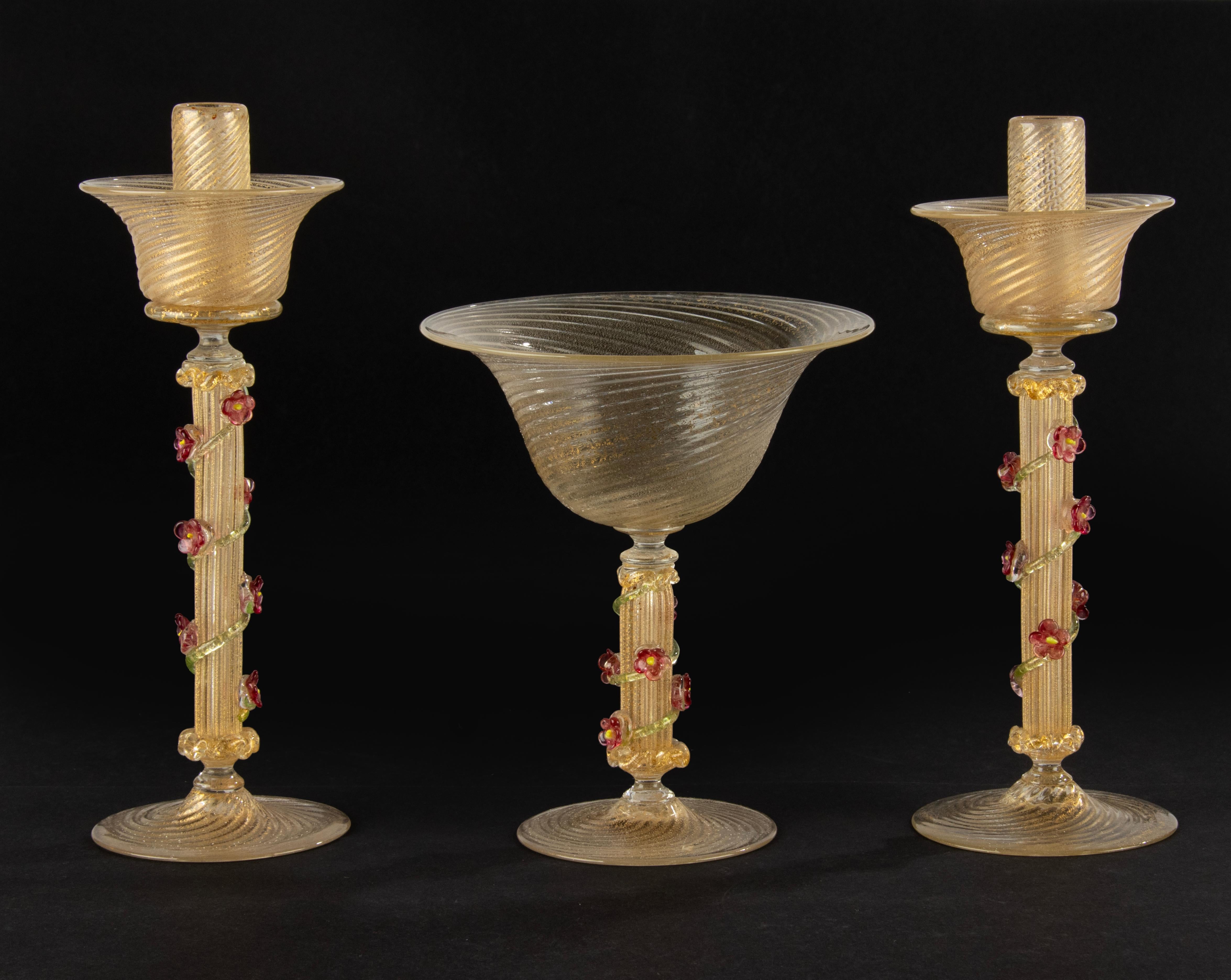 A beautiful set of 2 Murano glass candlesticks and a bowl. 
The glass has beautiful gold colored inclusions, the stems are decorated with delicate flowers. 
Dimensions: the candles are 26 cm tall and Ø9 cm, the bowl is 18 cm tall and Ø15 cm. 
All