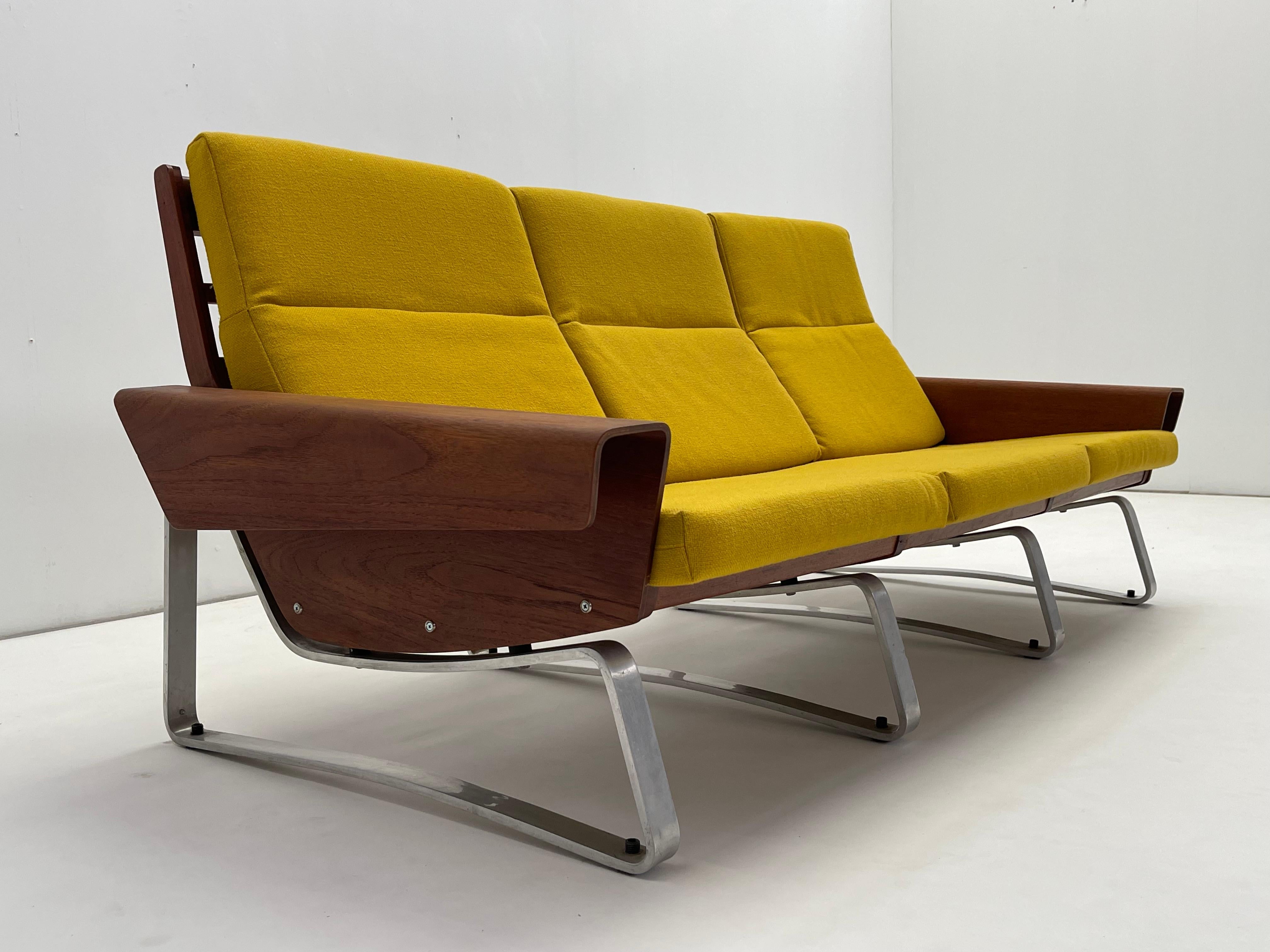 Questo splendido divano a 3 posti è un mix straordinario di design vintage e moderno, caratterizzato da un'elegante struttura in alluminio e da eleganti accenti in legno di teak. Il divano vanta linee audaci e angolari, con braccioli lisci e curvi