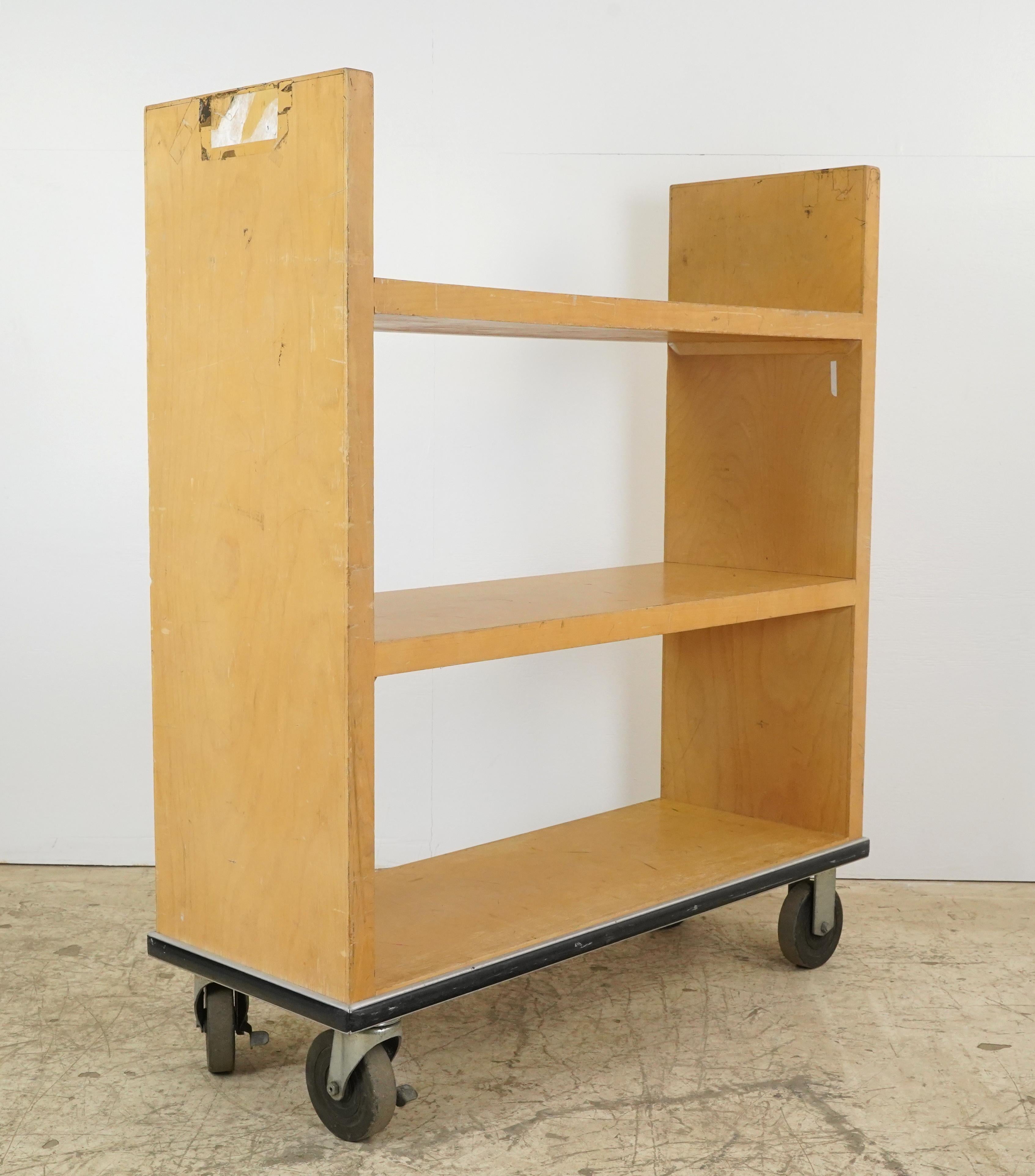 Reclaimed light maple industrial library book cart with three shelves and four rubber and steel casters. It has a simple functional design dating back to the 1970s. The cart features a black bumper around the base to protect it from damage. Good