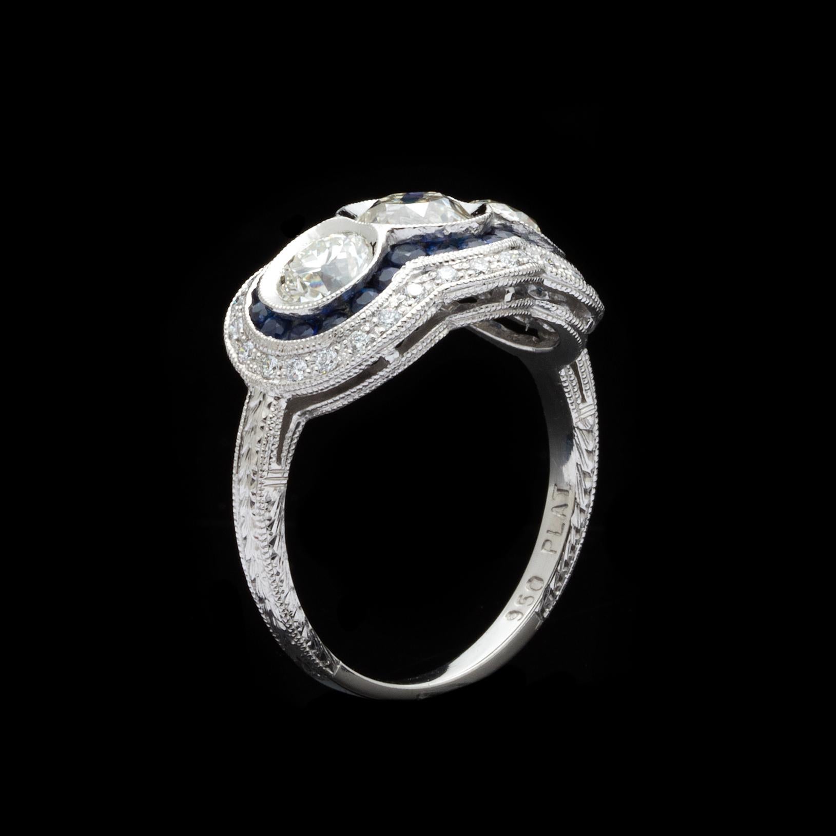 Platinum Edwardian style ring features 3 Old Euro cut diamonds totaling 1.14 carats. These diamonds are estimated as J/K color, and VS/SI clarity. Bordering the center stones is a halo of round cut blue sapphires totaling 0.48 carats, and 0.18