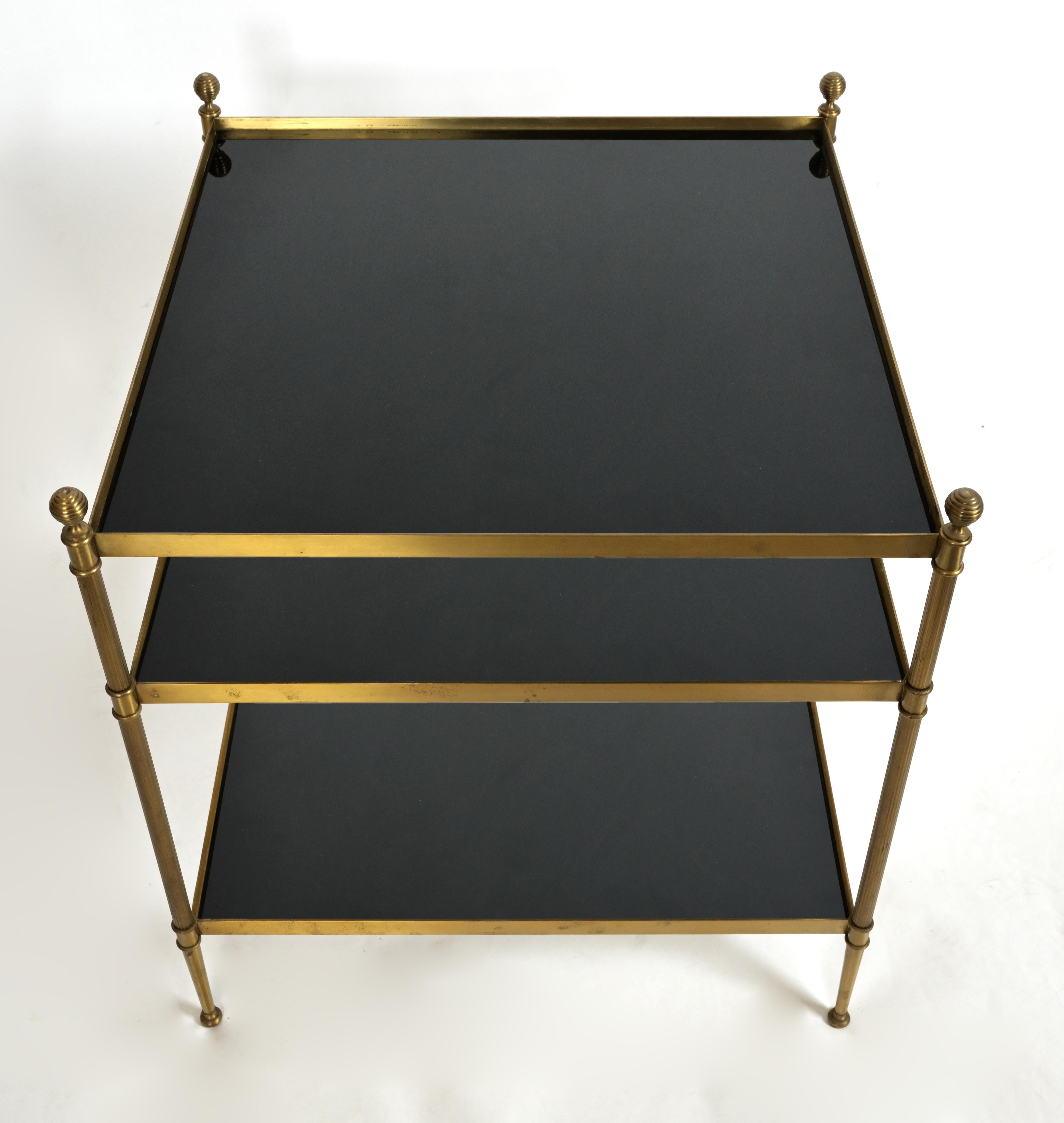 A square three tiered patinated brass side table with black vitrolite surfaces. The frame has furrowed finials capping the reeded legs at each corner. Designed in the manner of Maison Jansen, circa 1950.