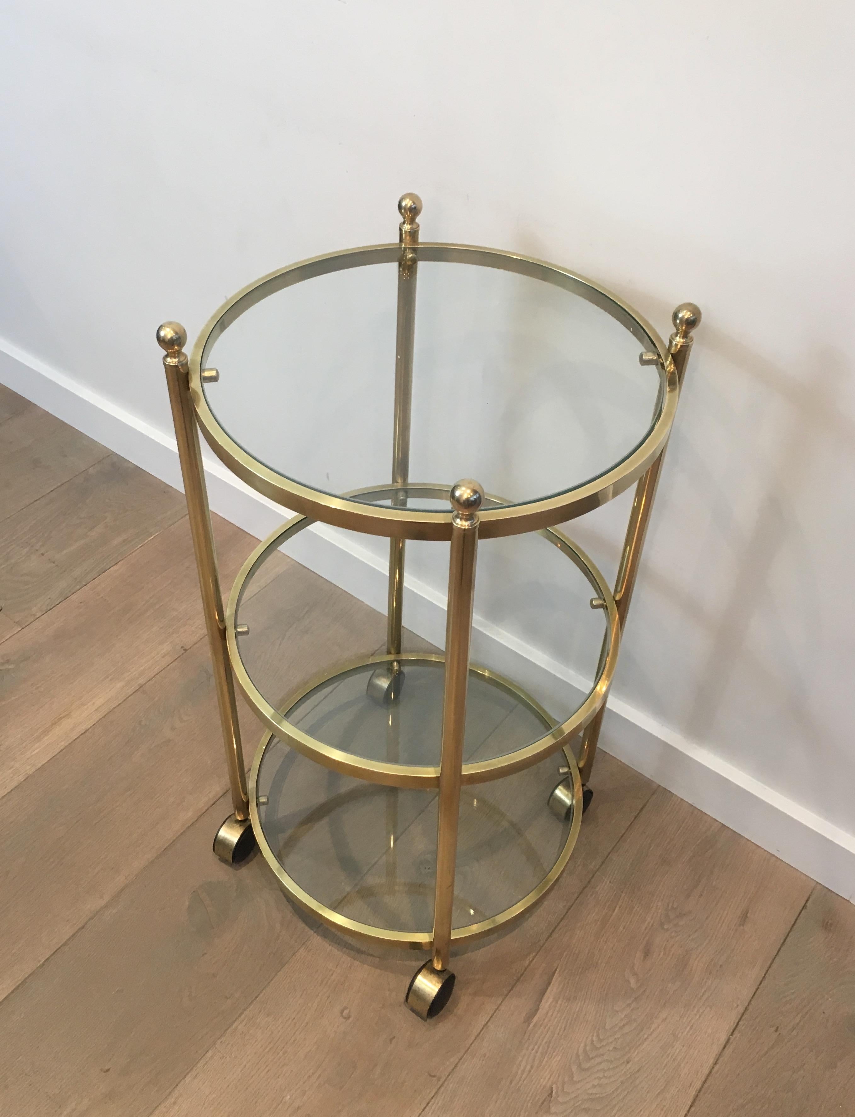 This 3 tiers round side table on casters is made of brass with 3 clear glass shelves and gilt and black plastic casters. The quality is very good. This is a French work, circa 1970.