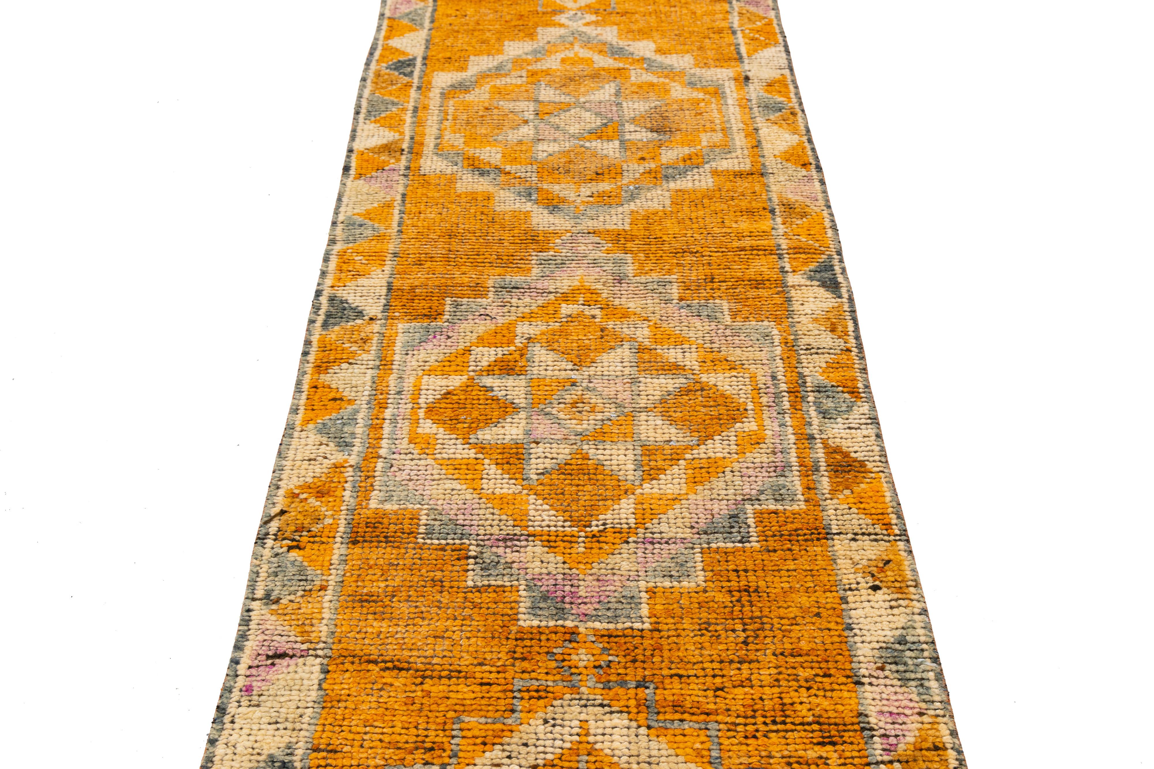 Tejida a mano en Turquía, esta vibrante alfombra presenta tres medallones centrales en forma de diamante rodeados de intrincados motivos tribales en cálidos tonos naranja, rosa y beige. Elaborado con lana natural, su diseño simétrico y su atrevida