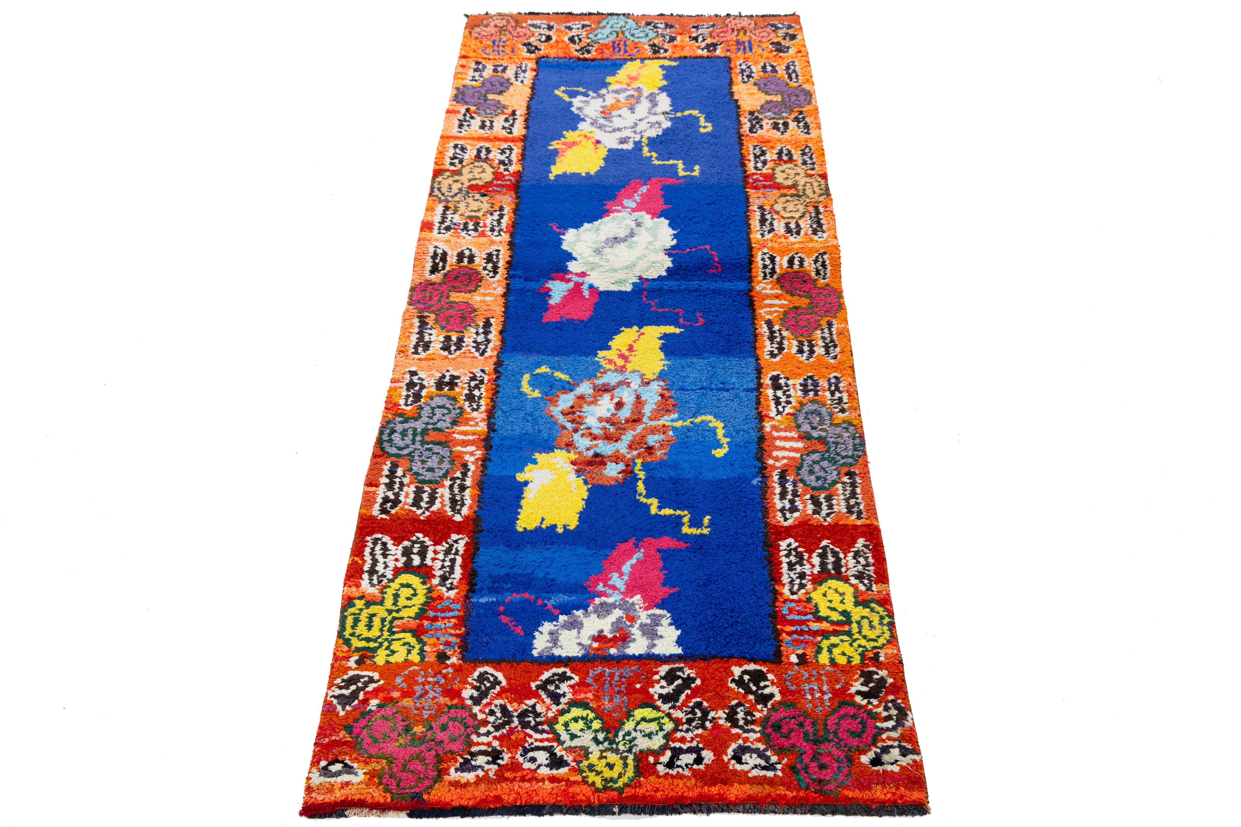 Dieser auffällige, in der Türkei handgewebte Läufer zeigt ein tiefblaues Feld mit wirbelnden, mehrfarbigen abstrakten Formen, die von einer lebhaften orangefarbenen Bordüre mit stilisierten Blumenelementen eingerahmt werden. Inspiriert von der