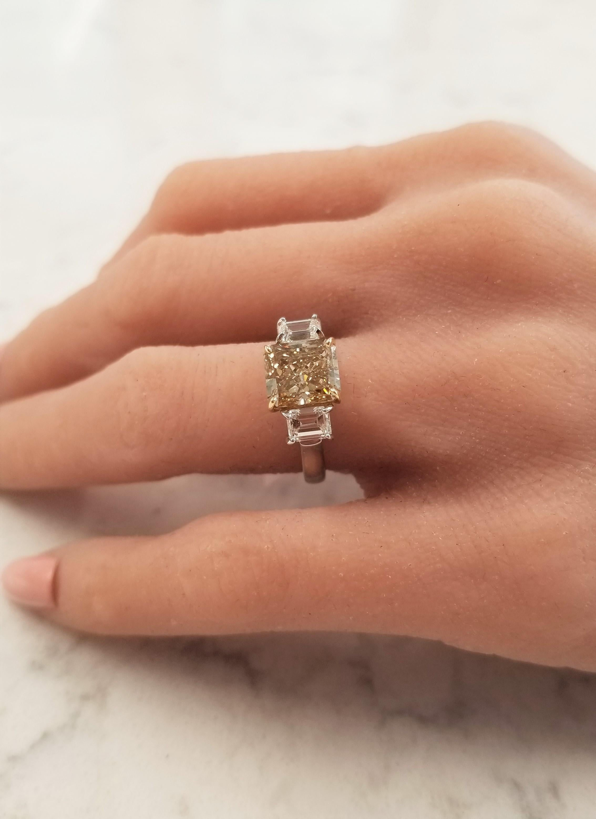 This enchanting three-stone diamond ring features a 3.00 carat radiant cut, natural fancy brown-yellow, VS2 clarity, diamond in the center and measures 8.3X7.4mm. Its evenly distributed color is a mix of cinnamon and honey. It is accentuated by rich