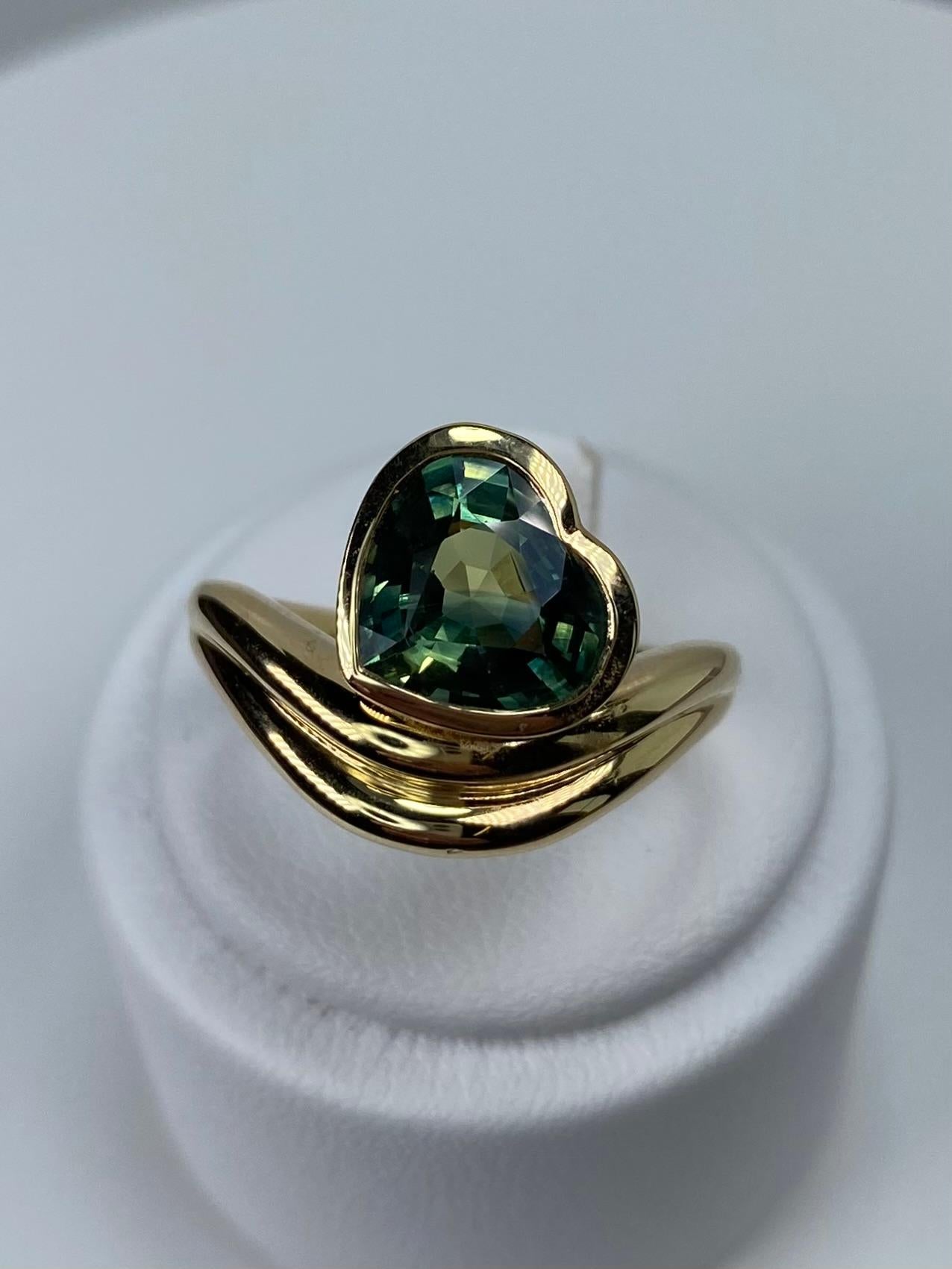 Introducing an exquisite masterpiece from The Sapphire Merchant: this 18k yellow gold and 3.00ct heart-shaped teal sapphire ring. This captivating ring boasts a unique rub-over setting that elegantly encases the enchanting greenish teal sapphire,