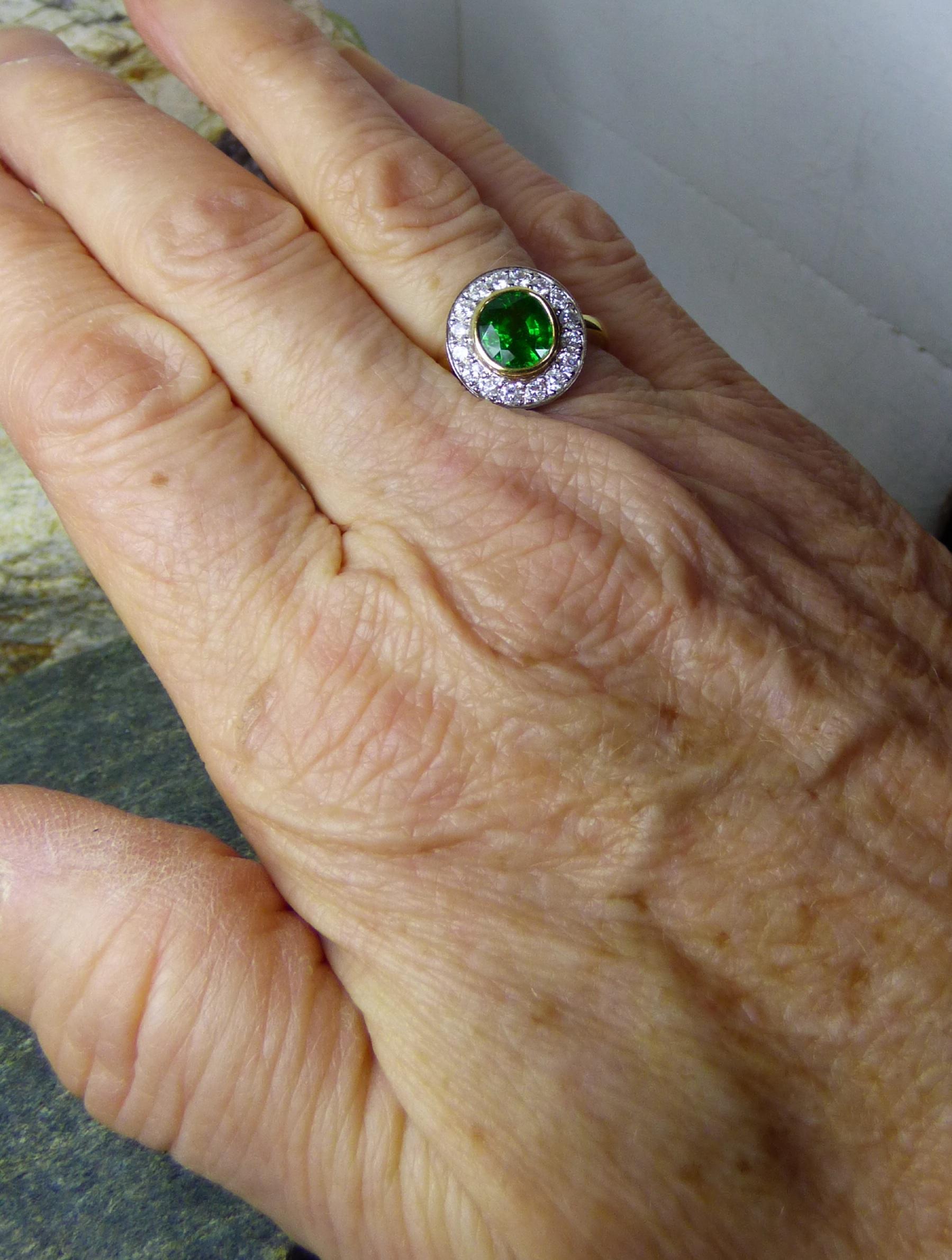 A delightfully bright and colourful Tsavorite Garnet oval stone is 7X6mm and weighs 3.01ct. The Garnet is surrounded by 16 Diamonds with a total weight of .76ct.  The ring is handmade in 18k yellow gold with the Diamonds set in 18K white gold.  The