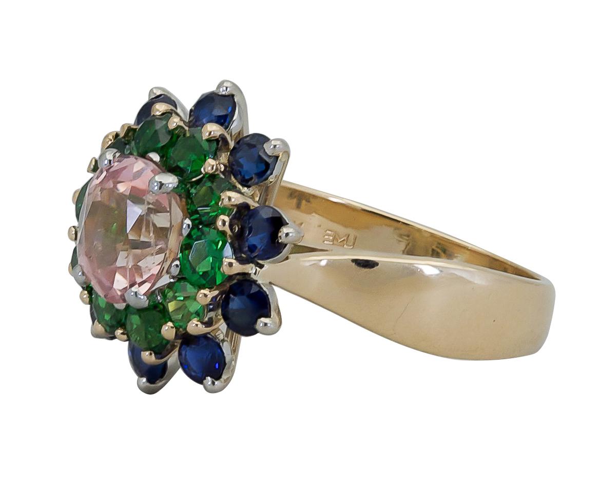 A fashionable ring showcasing an oval cut pinkish sapphire, surrounded by a row of vibrant round tsavorites and blue sapphires. Set in a polished 18k yellow gold mounting.
Center stone weighs 3.02 carats.
Tsavorites and blue sapphires weigh 2.10