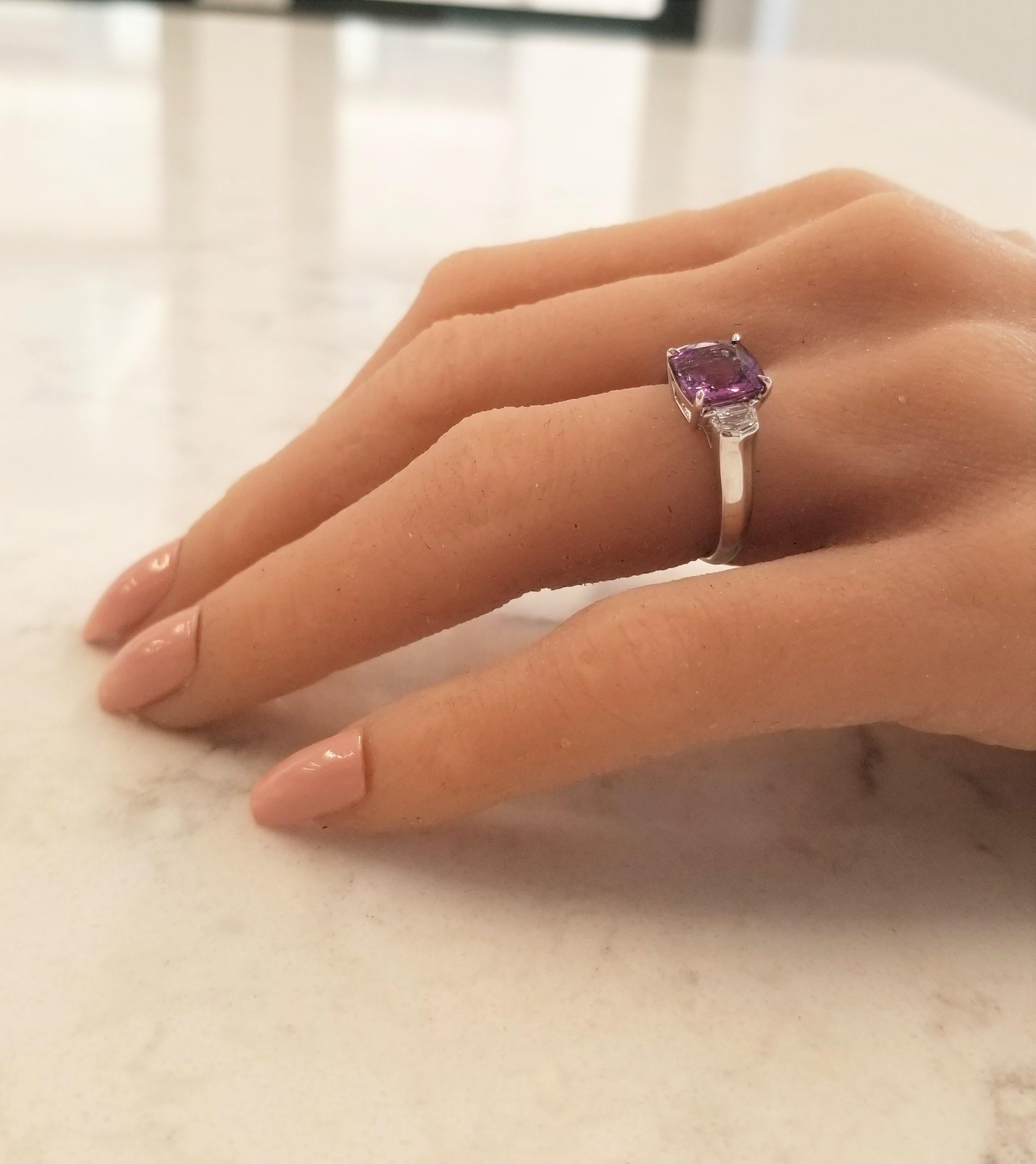 This is a 3.04 carat square cushion natural pink sapphire. The gem source is Sri Lanka. Its color is vivid purplish pink. Its transparency and luster are excellent. Two cadillac-cut white diamonds hug this dynamic sapphire totaling 0.48 carats, all