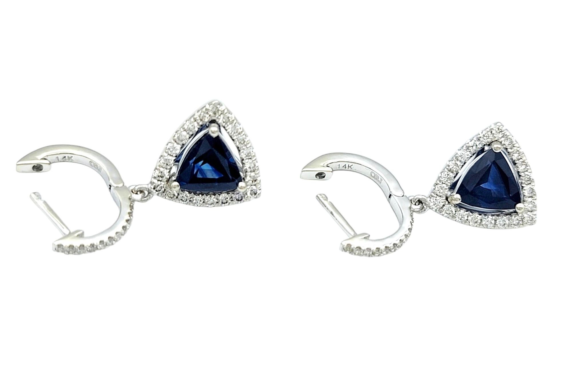 This stunning pair of drop dangle earrings, set in elegant 14 karat white gold, is a stunning display of sophistication. Each earring features a triangular blue sapphire at the center, surrounded by a halo of round diamonds. The combination of the
