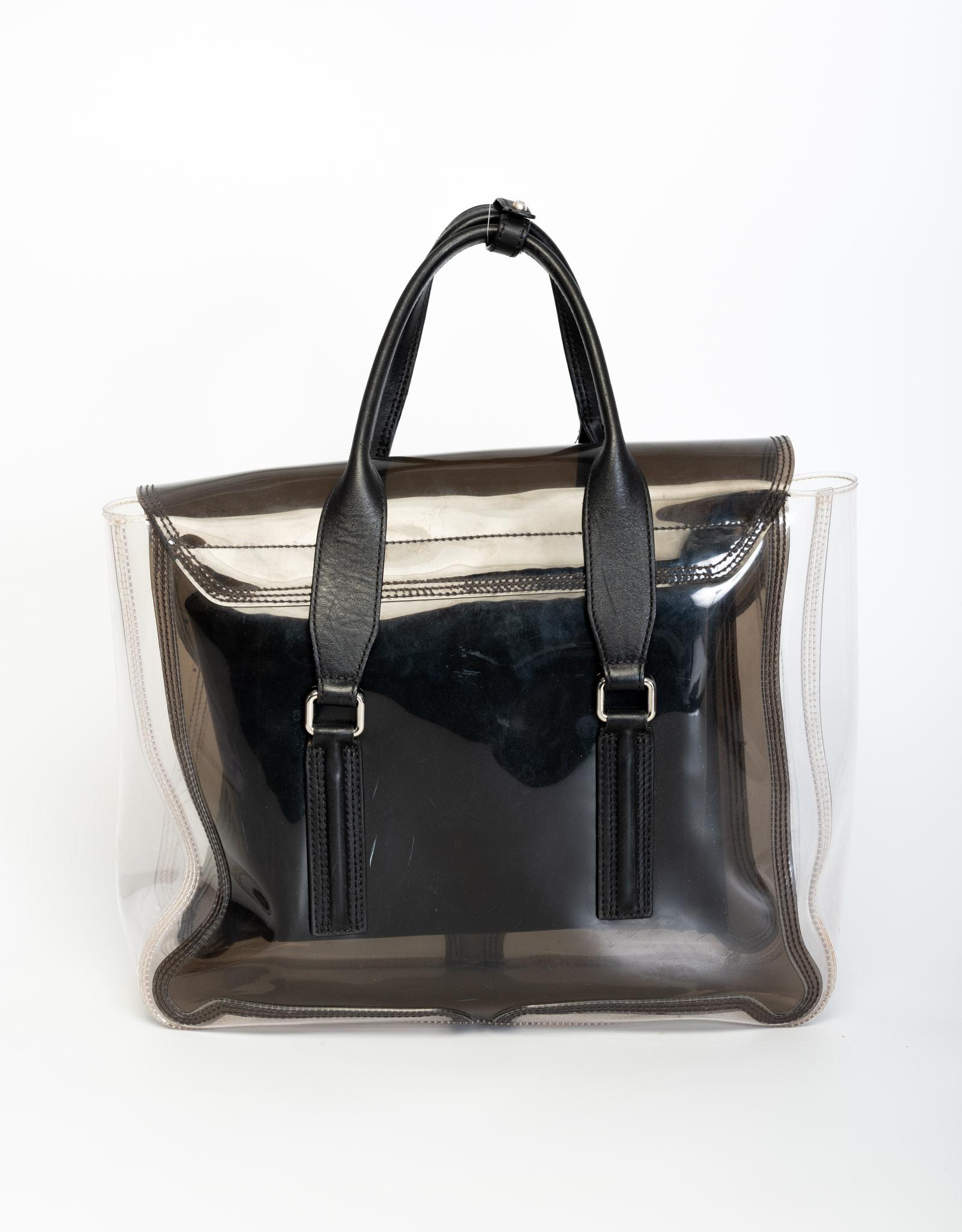 This transparent plastic satchel features a front flap with two expandable vertical zippers beneath it. Featuring leather top handles, silver tone buckle closure and an internal leather zipper pocket.

COLOR: clear/smoky black
MATERIAL: PVC with