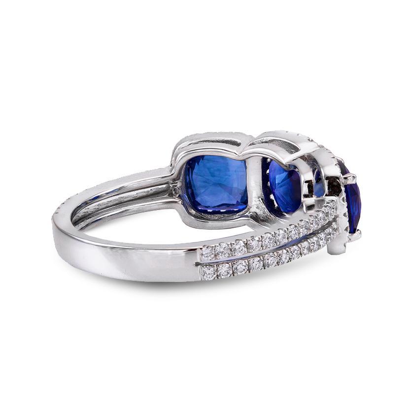 Un anillo de compromiso a la moda, con tres zafiros azules naturales talla cojín de 3,25 quilates en total, cada uno engastado con cuatro garras. Rodeada por un halo compartido de diamantes redondos de talla brillante y acentuada por un cuerpo de