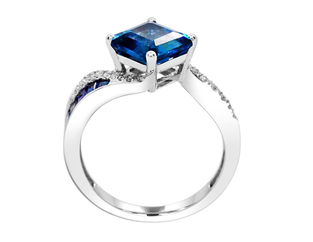 This cushion cut London Blue Topaz, Sapphire and Diamond ring is set in 14K White gold.  This ring has a 3.26 carats total weight of London Blue Topaz set at the center, with baguette of varying sizes weighing 0.89 carat total weight.  The 8 x 8 mm