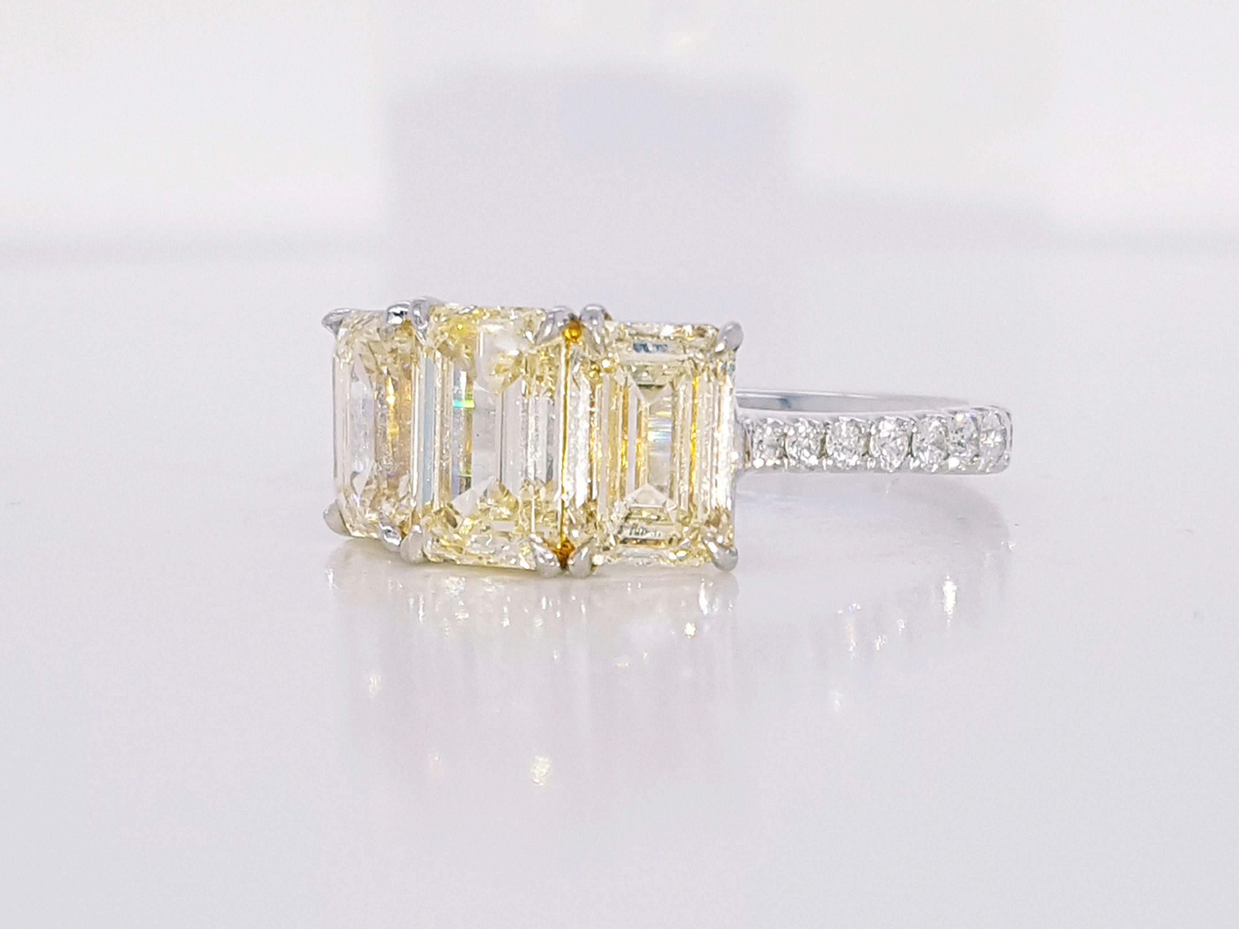 This gorgeous engagement ring features an emerald cut yellow diamond prong set in the center of a three-stone setting, showcasing a 3.35 Carat Yellow Diamond Emerald Cut Three-Stone Engagement Ring, accented by 18 round brilliant diamonds G color,