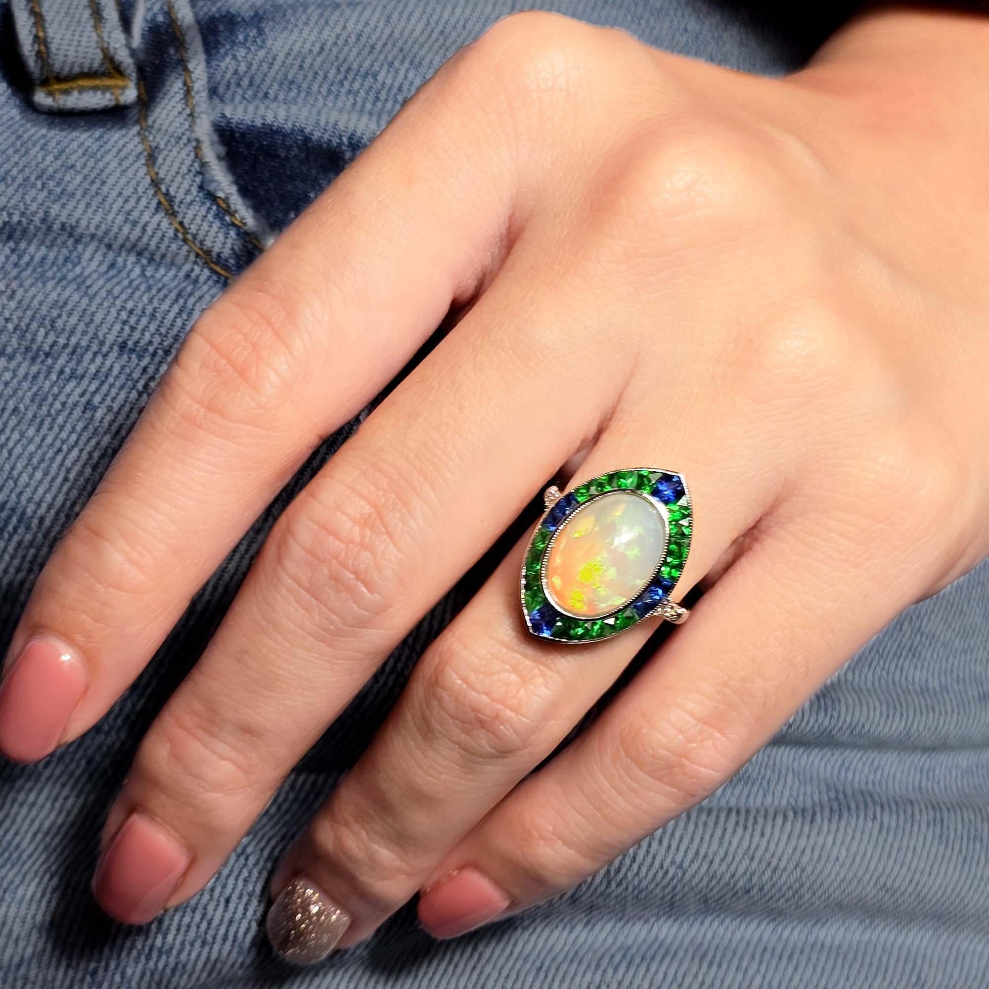 L'opale éthiopienne de 3,5 carats au centre, avec son jeu de couleurs envoûtant. À chacune des quatre pointes, des tsavorites vibrantes sont serties, ajoutant un riche contraste vert. Autour de l'opale, un anneau de saphirs étincelants rehausse