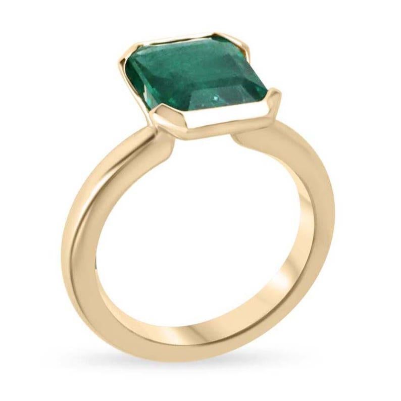 This captivating solitaire ring features an emerald-cut emerald set north to south, showcasing a vivid deep green color with mesmerizing semi-transparent clarity. The emerald is nestled in a distinctive half-bezel setting, giving the piece a bold,
