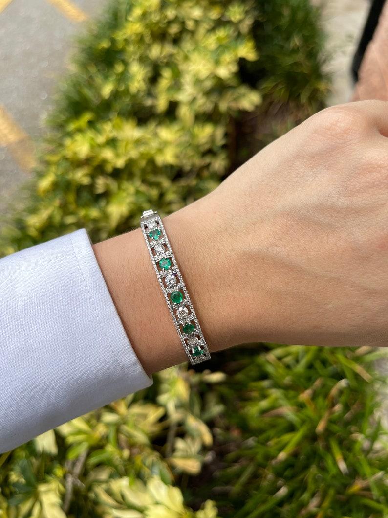 Adorn your wrist with the exquisite beauty of our emerald and diamond bangle bracelet, featuring five round-cut emeralds of medium rich green color and semi-transparent clarity. Brilliant round-cut diamonds, totaling 2.50 carats, accentuate the