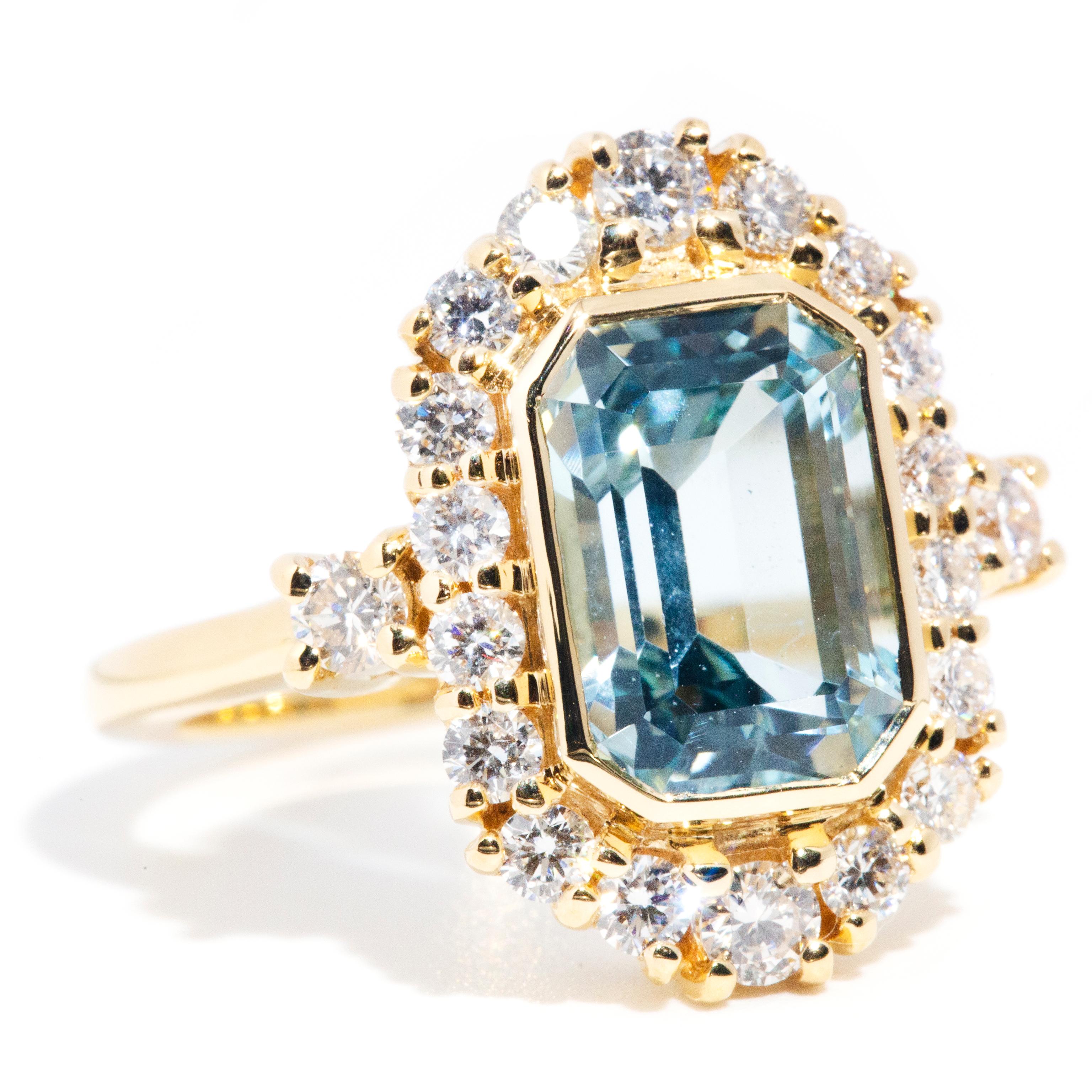 Crafted in 18 carat yellow gold, this stunning contemporary ring is breathtaking with her wonderous emerald cut aquamarine glowing in the centre and encompassed by a surrounding border of radiant round brilliant cut diamonds. This masterwork is