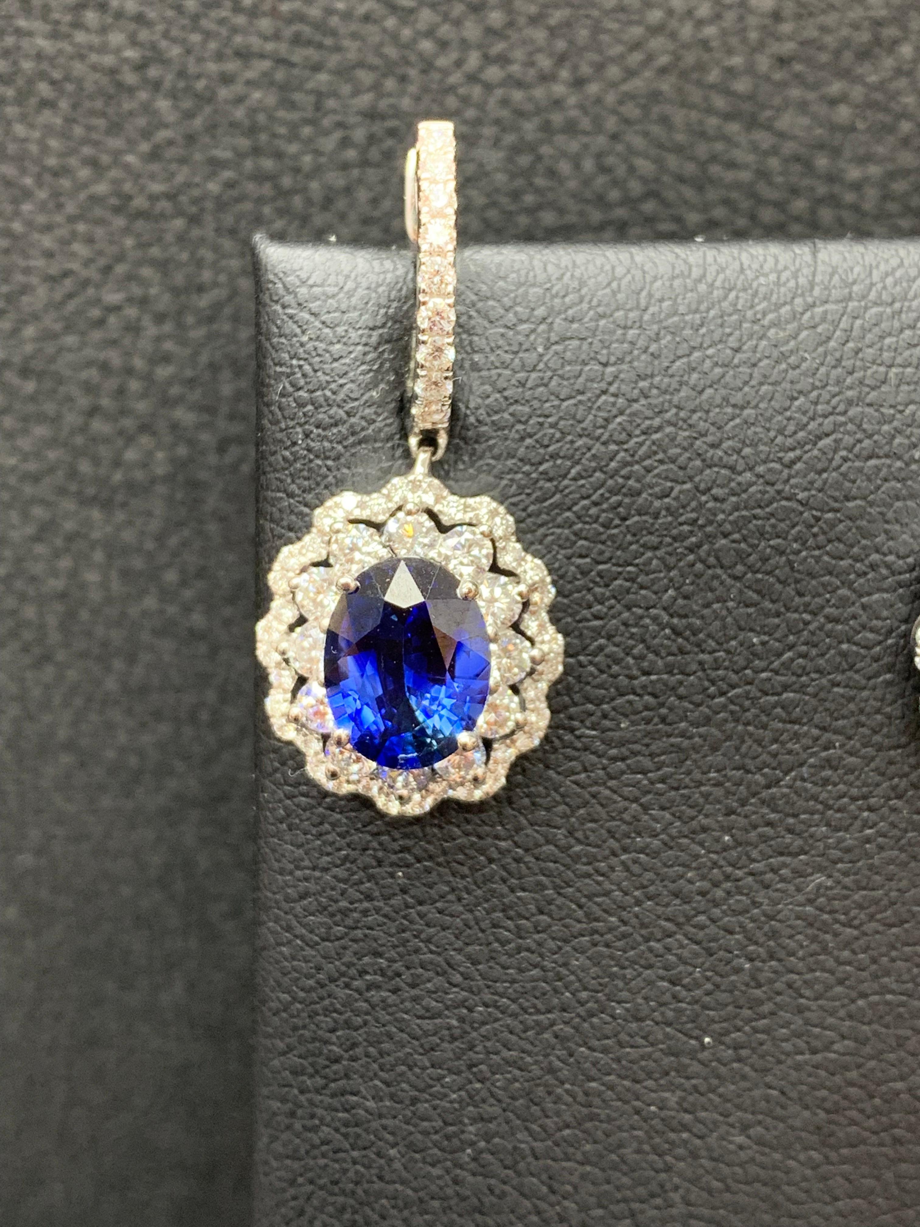Dangle earrings featuring Oval shape blue sapphires weighing 3.56 carats total, surrounded by two rows of round brilliant diamonds in a flower design. Accent Diamonds weigh 1.54 carats total. Made in 18 karat white gold.